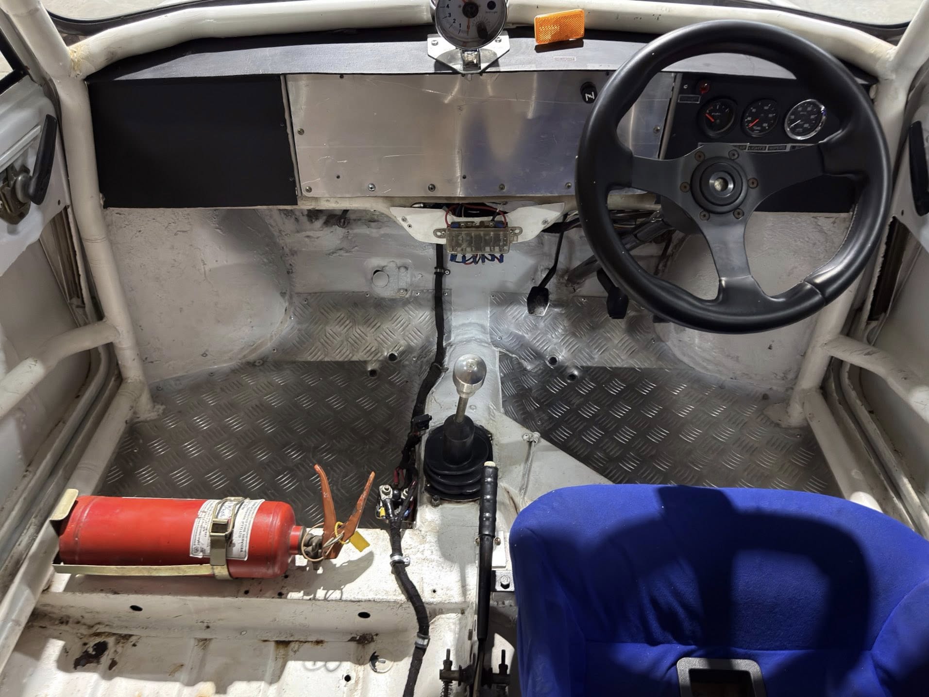 Racing car cockpit with steering wheel on right, metal dashboard, gear lever, and blue racing seat; fire extinguisher mounted on left.