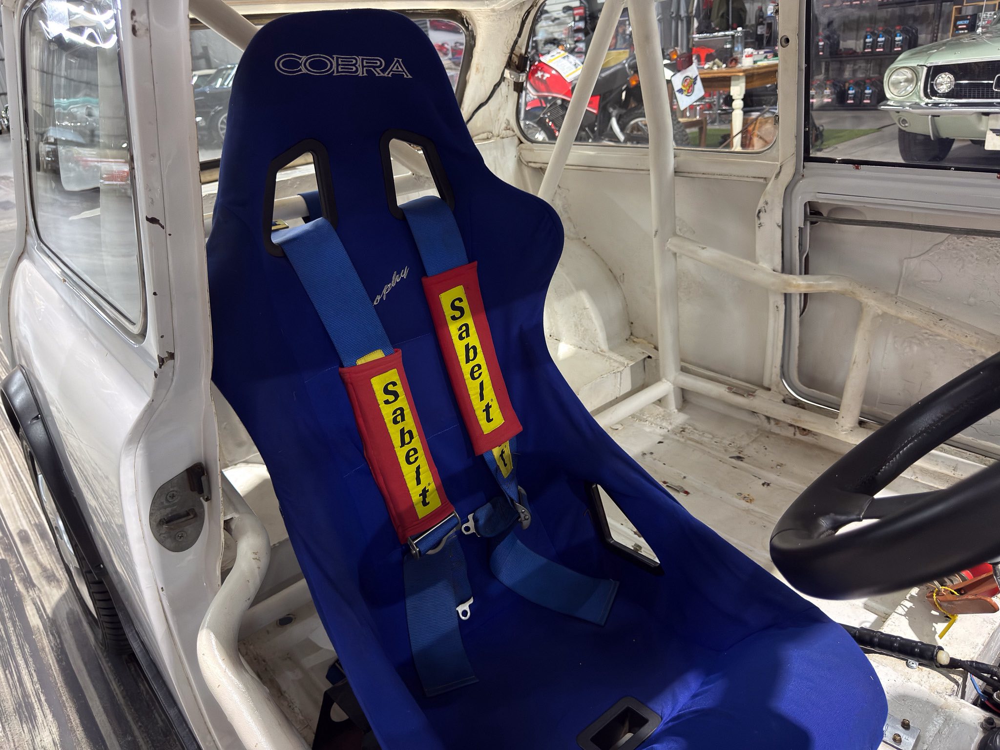Blue Cobra racing seat with a blue five-point harness inside a white race car, exposed roll cage visible.