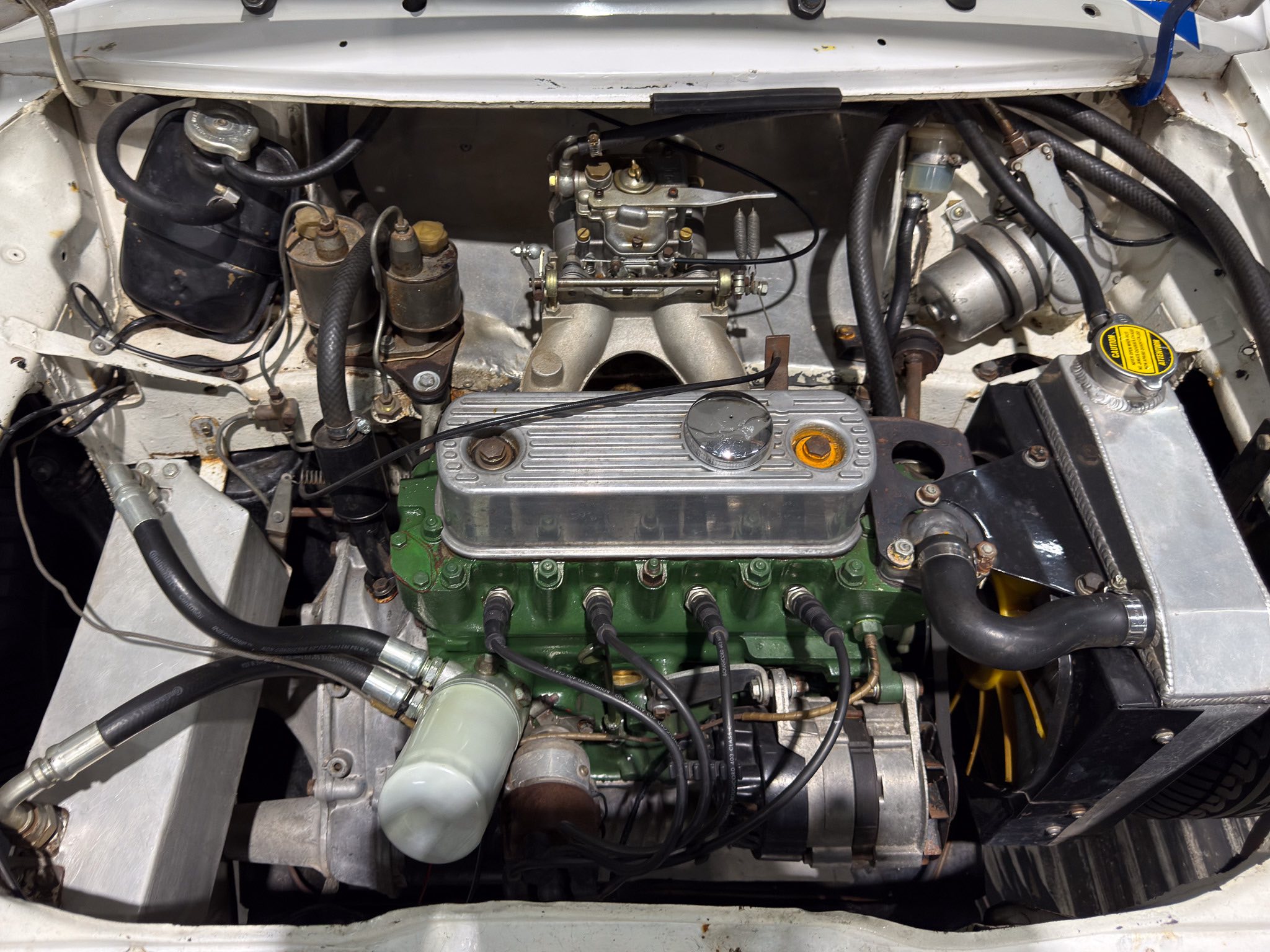 Open engine bay of a vintage car showing a carburetor, intake manifold, hoses, and a green engine block visible below the valve cover.