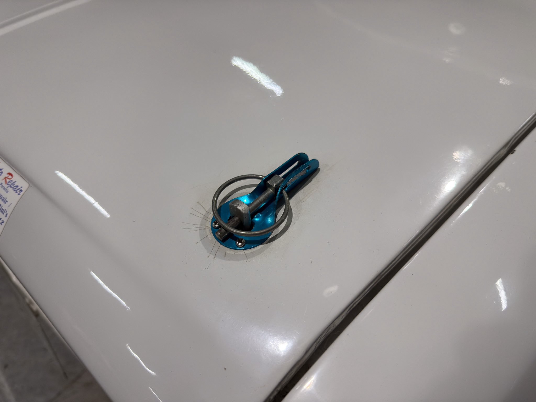 Blue automotive dent-removal tool rests on the light gray car hood, with a black cord nearby and metal hardware visible.