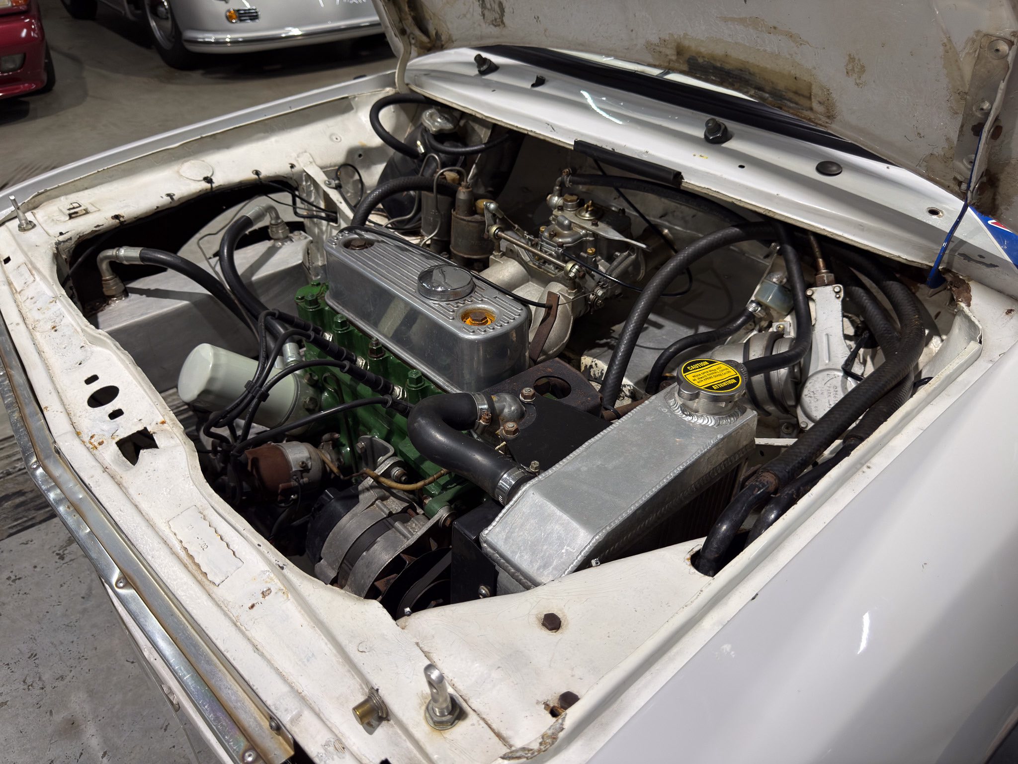 Open hood of a classic car showing the engine bay with a chrome valve cover, hoses, and radiator in a dusty workshop.