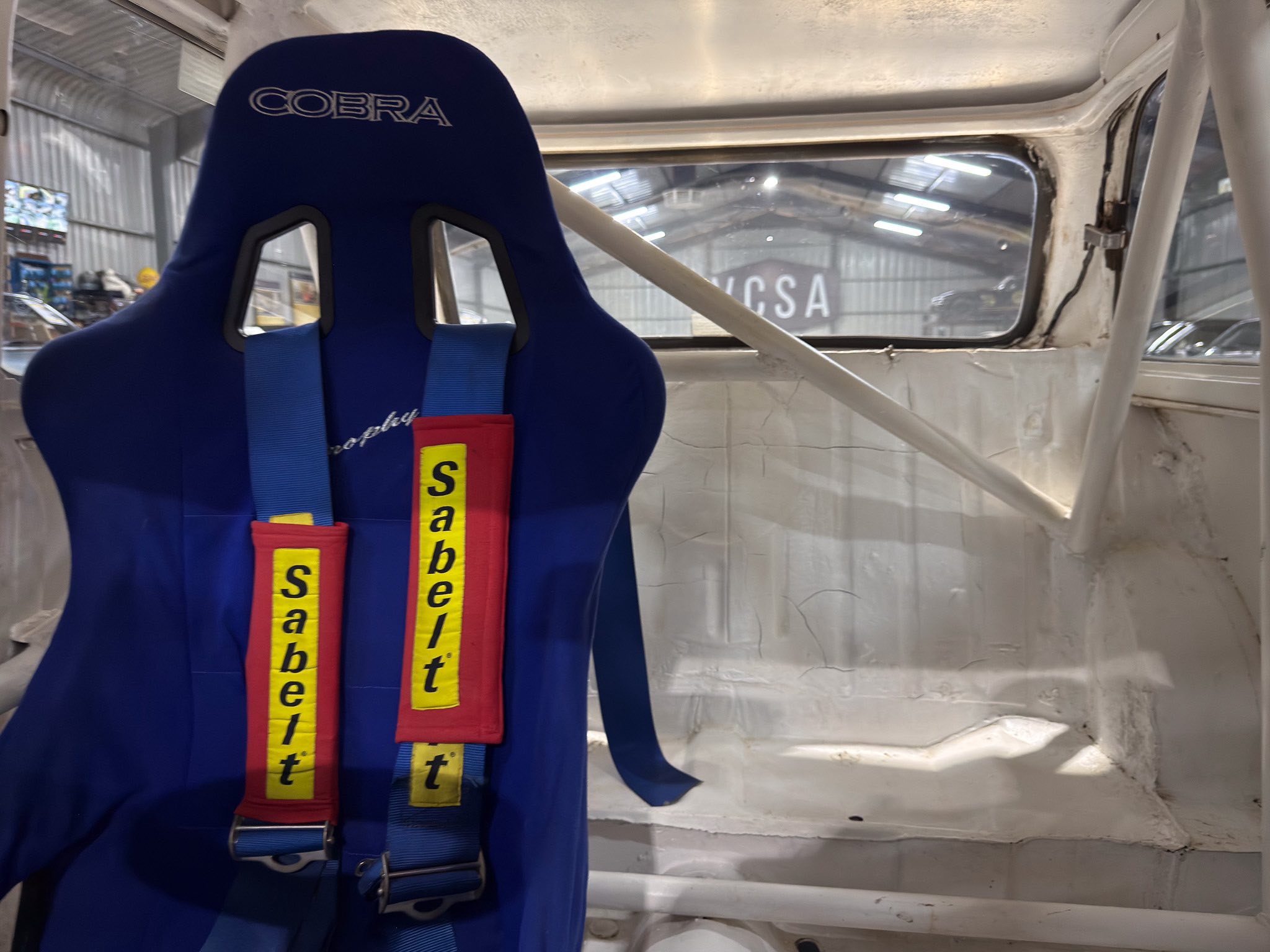 Blue racing seat with red/yellow Sabelt-style straps labeled 'seat belt' inside a workshop vehicle, COBRA embroidered on headrest.