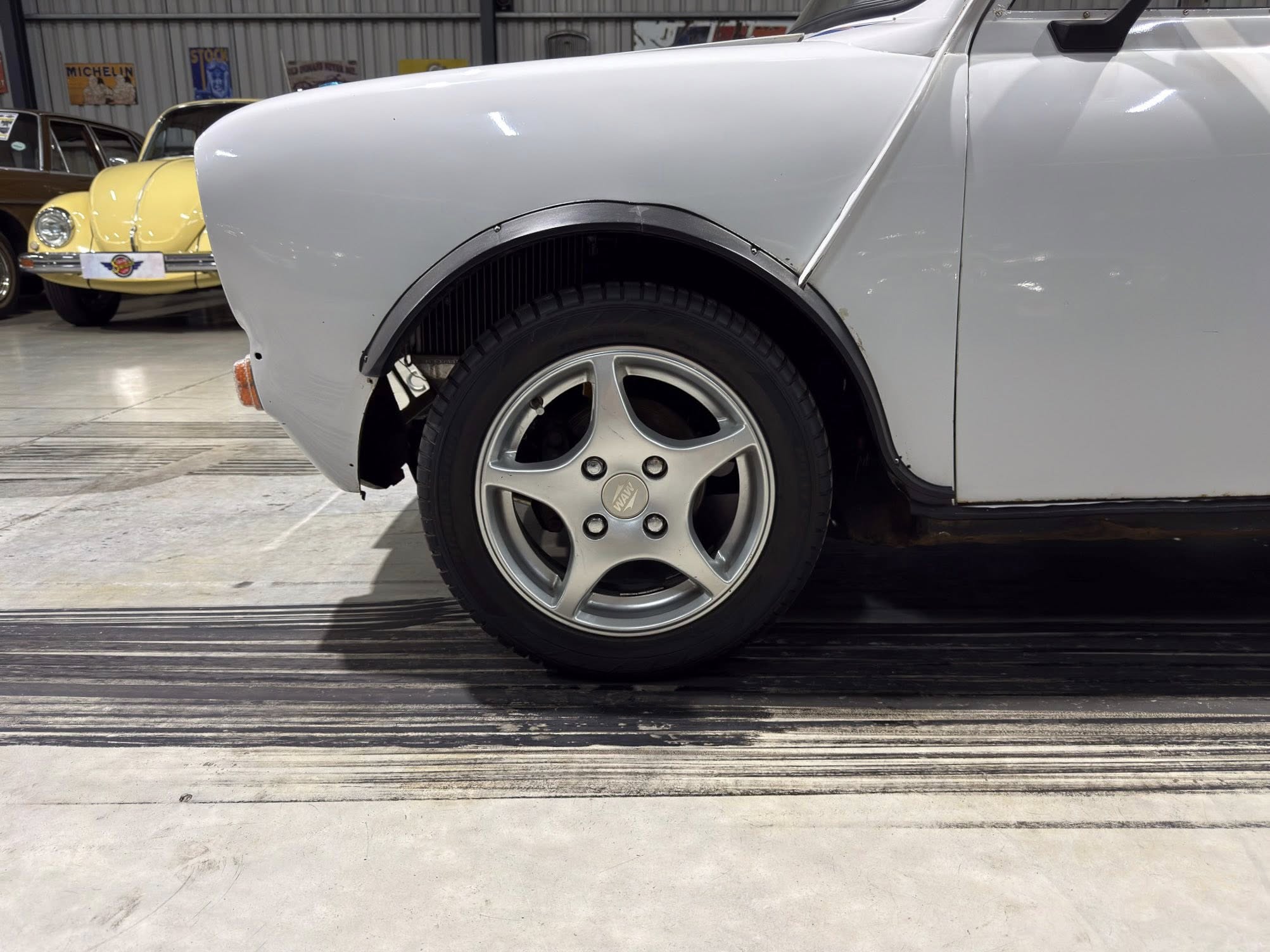 Close-up of a silver car's front left wheel and fender inside a showroom, with vintage cars in the background.
