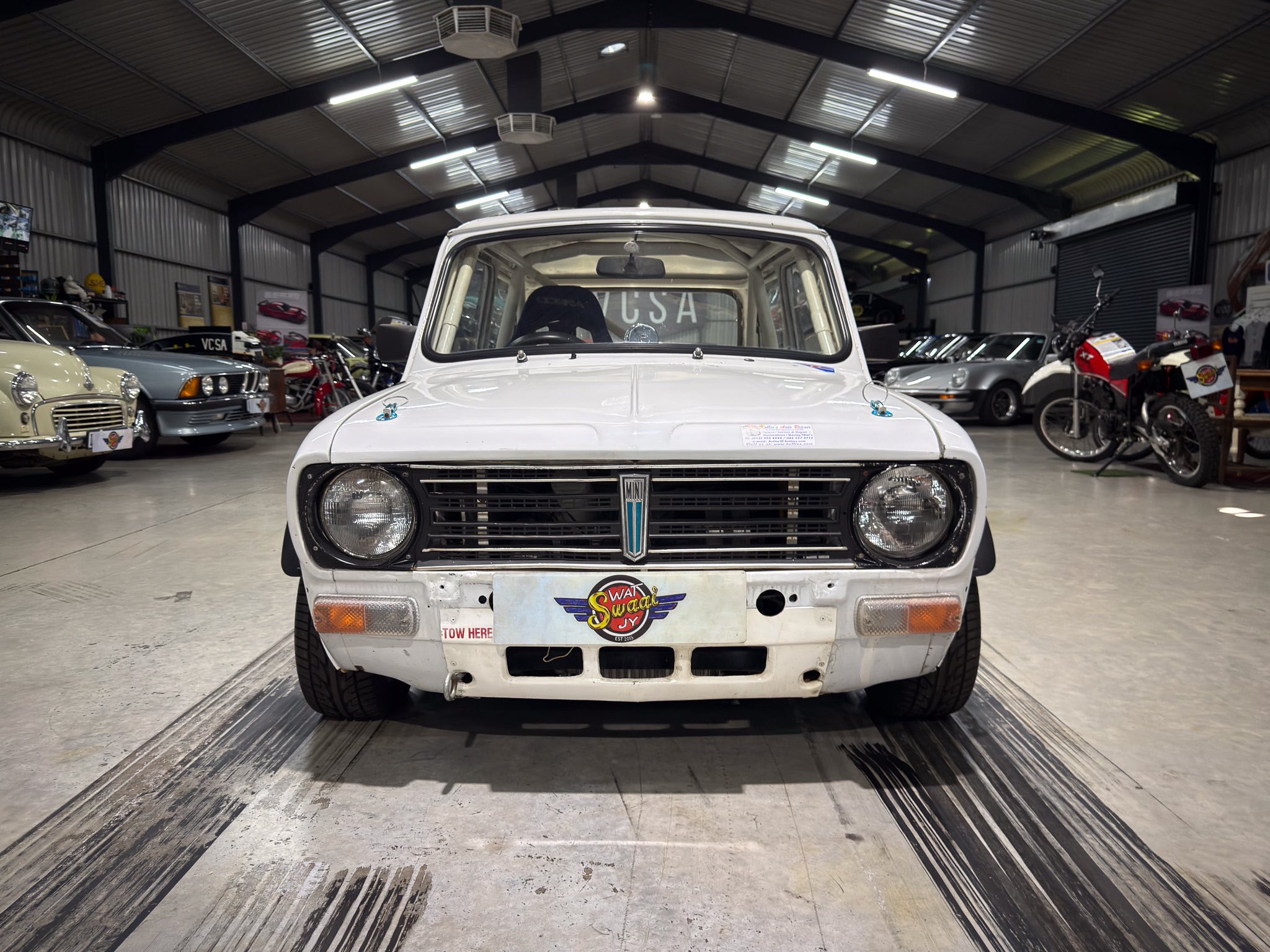 Front view of a white vintage car in a showroom, with other classic cars and motorcycles along the sides.