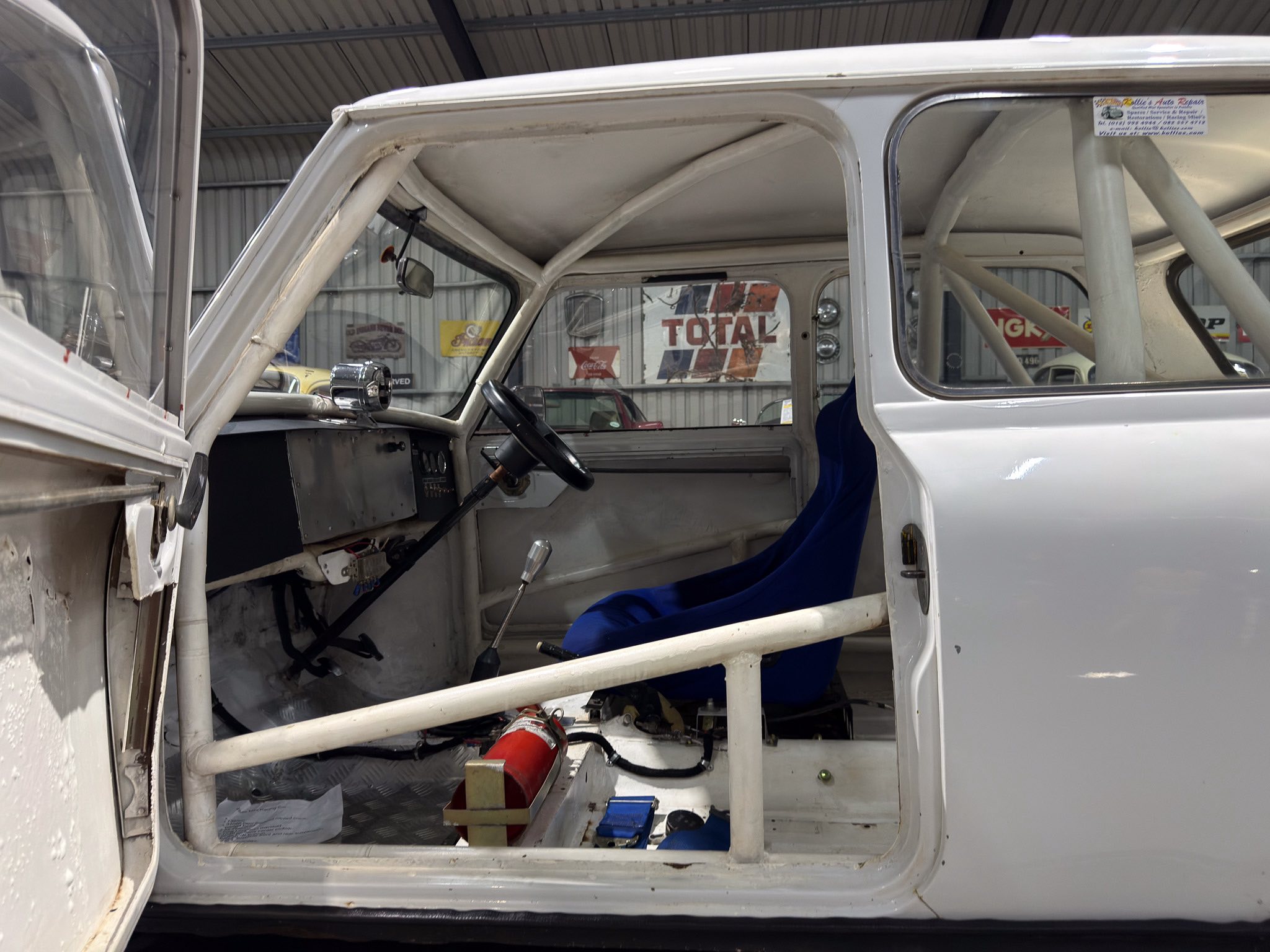 Interior of a stripped vintage white car with exposed roll cage, steering wheel, and blue seat cover; a red fire extinguisher sits on the floor amid exposed metal panels in a workshop garage.