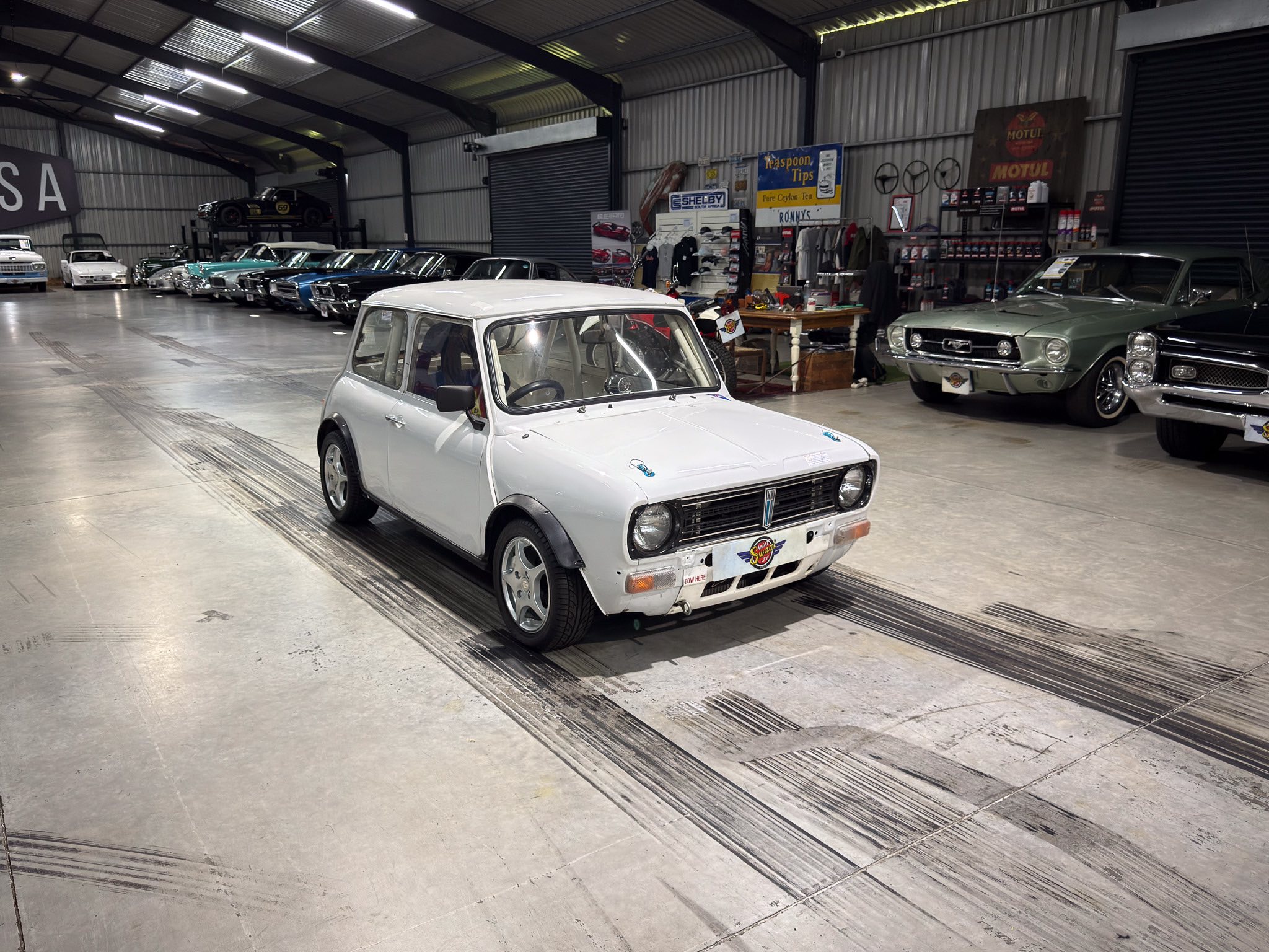 Indoor classic car showroom with a white compact car in the foreground and vintage cars lined up along the left wall, USA sign visible on the left side.
