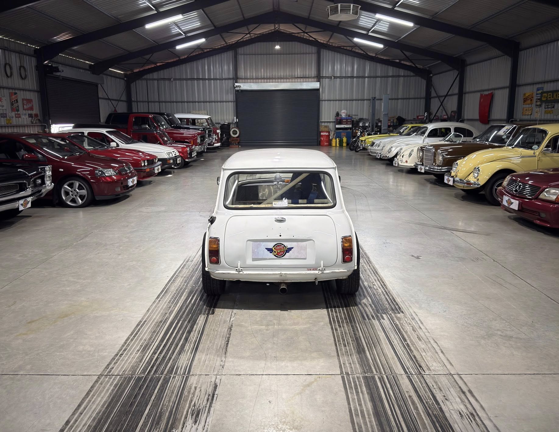 Rear view of a white vintage car in a spacious showroom lined with classic cars on both sides.