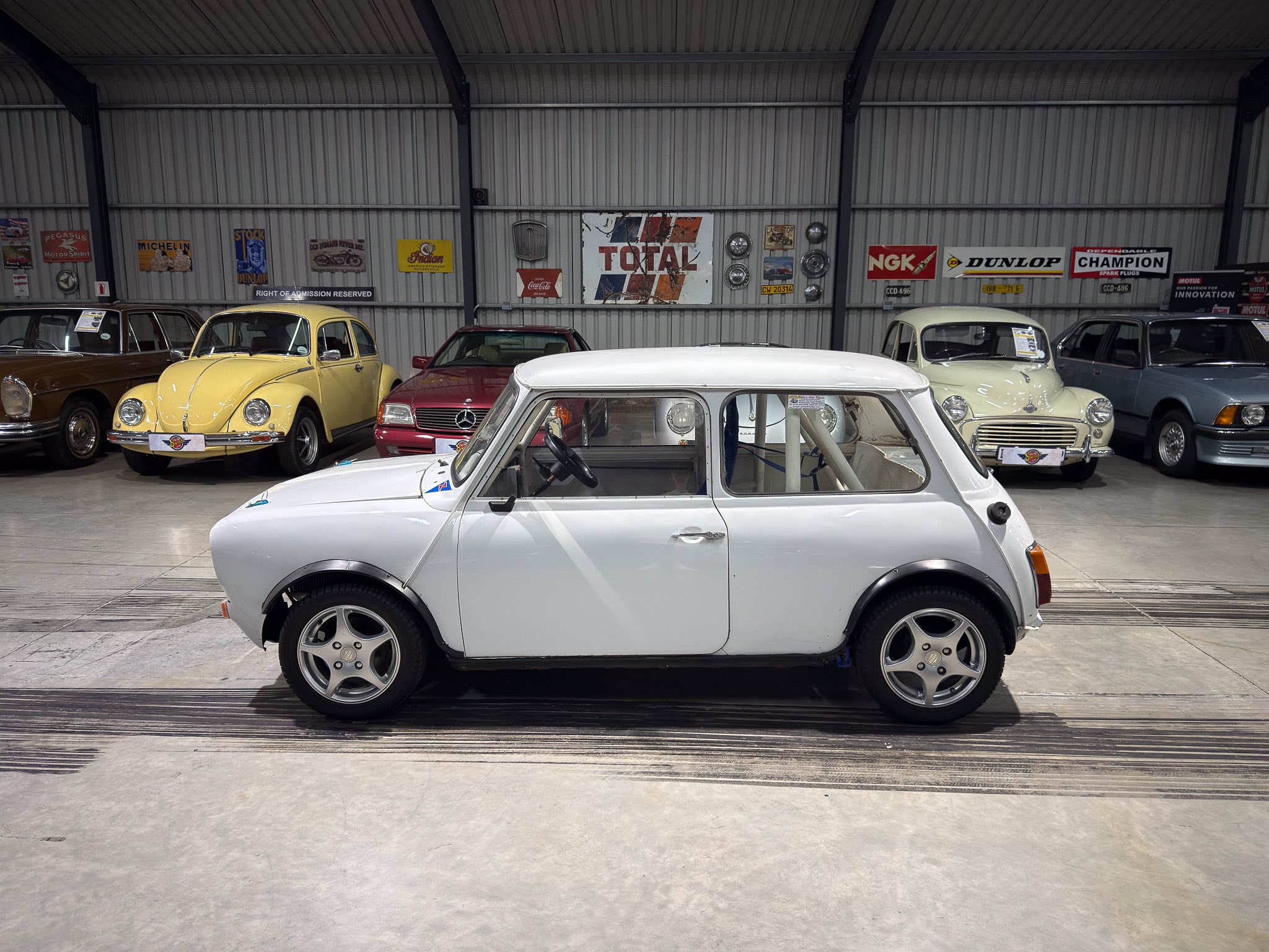 Vintage cars lined up inside a metal-walled showroom with retro signs on the wall behind them, including a yellow Beetle and a white car in the foreground.