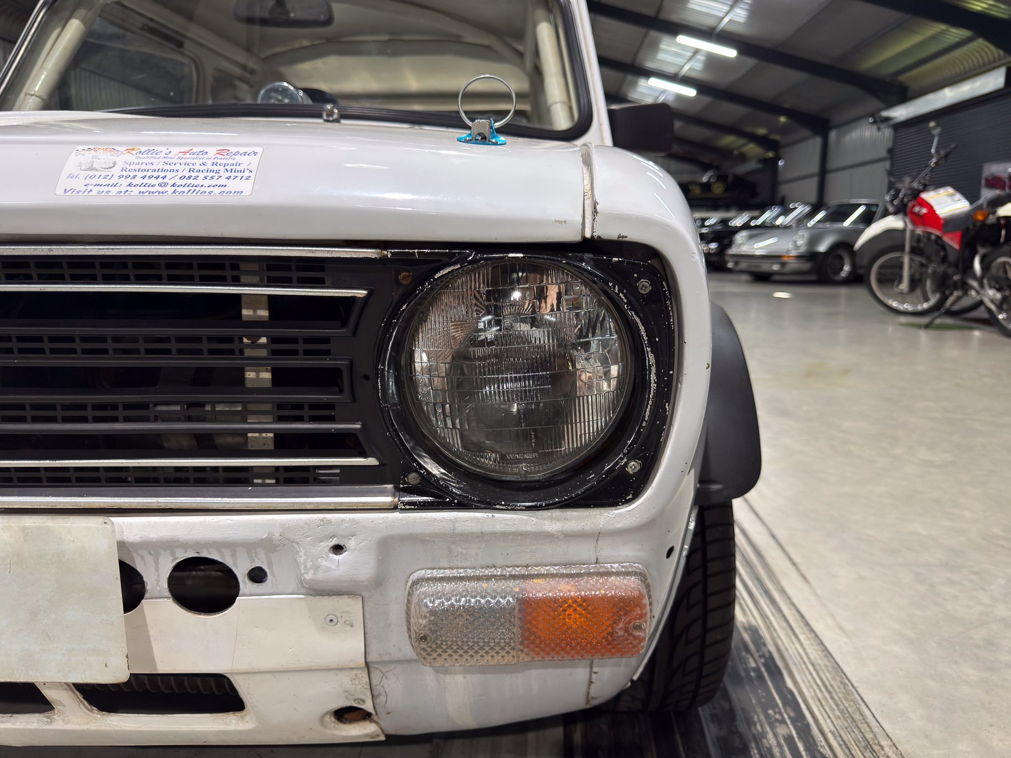 Close-up of the front left of a white vintage car in a garage; headlight, damaged bumper, and a blue-ring antenna on the hood.