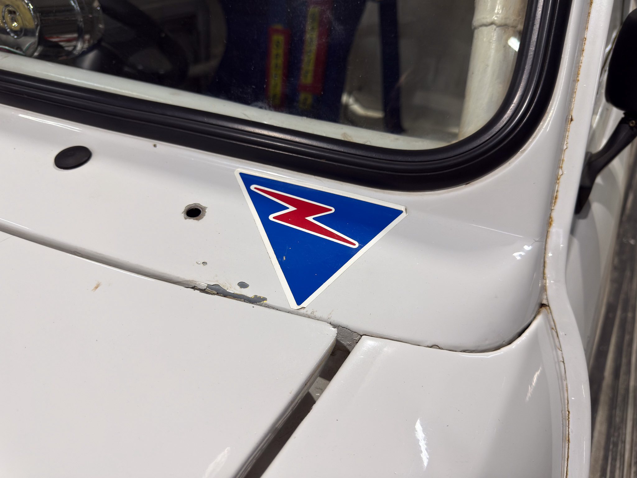 Close-up of a white vehicle body with a blue triangular badge, a black rubber window seal, and rust along the edge with chipped paint.