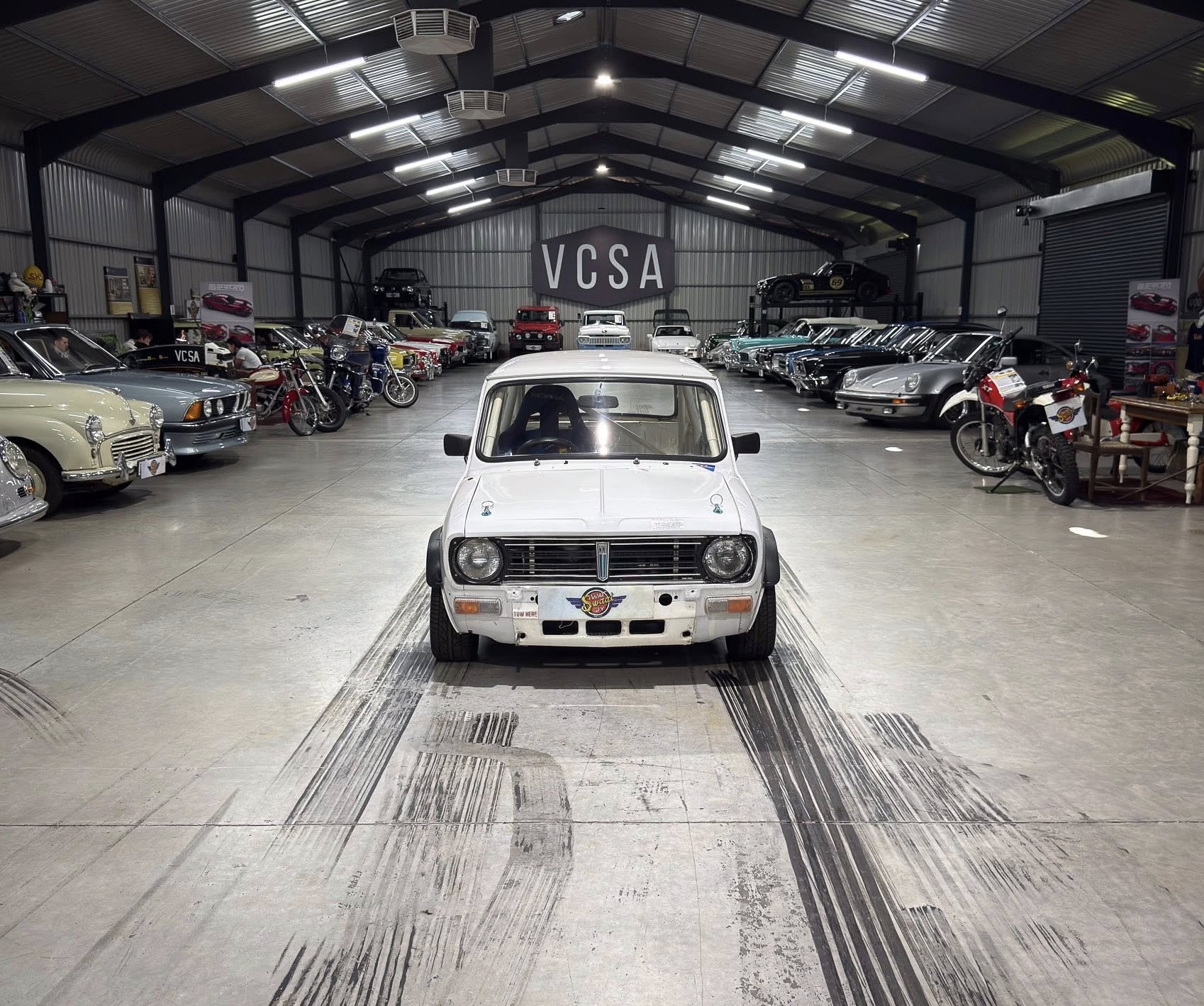 White vintage car centered in a large indoor showroom, with rows of classic cars and motorcycles on both sides and a VCSA sign in the back.