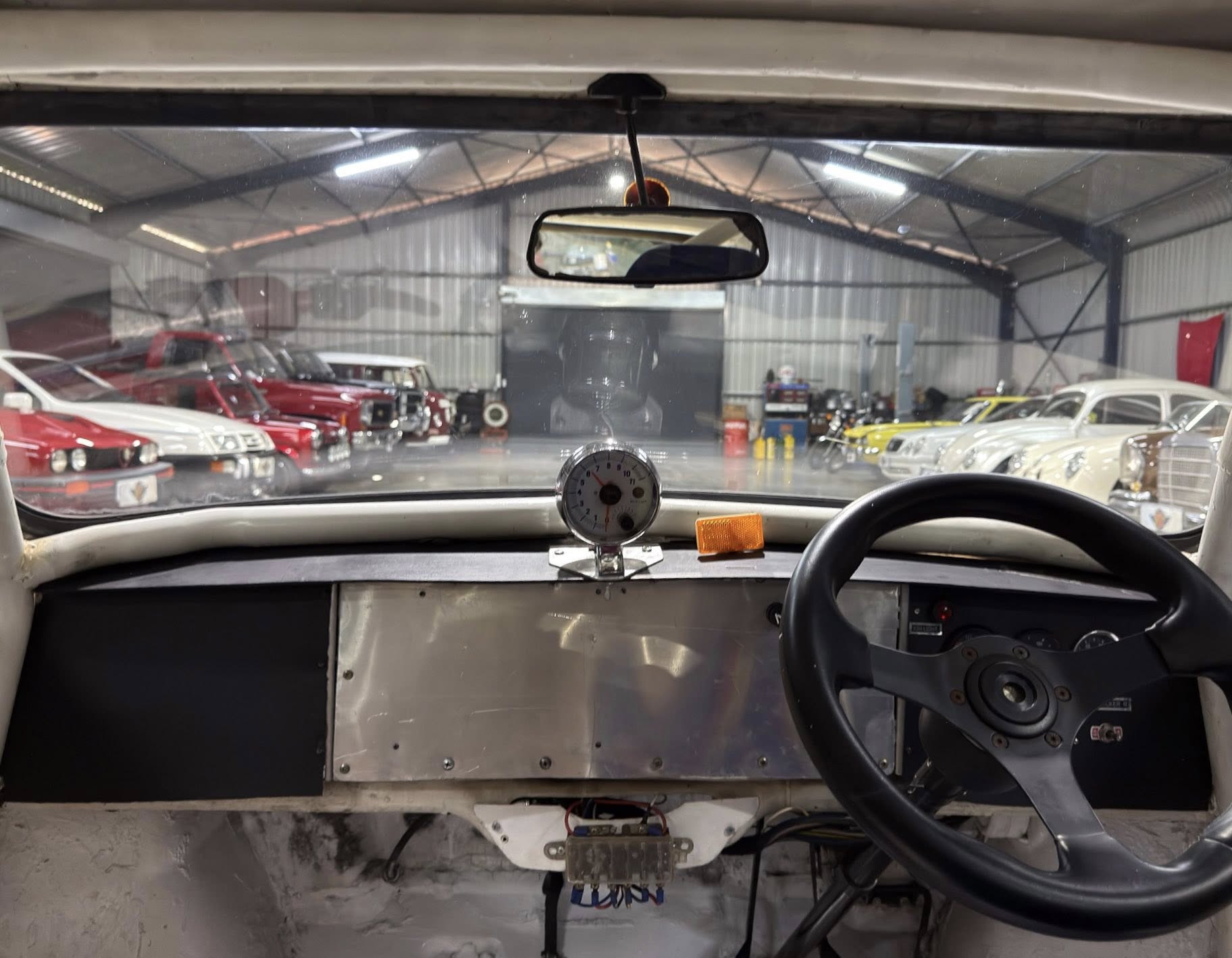 Cockpit view from a race car, showing a metal dashboard, steering wheel, and a round gauge, looking out into a workshop with classic cars parked along both sides.