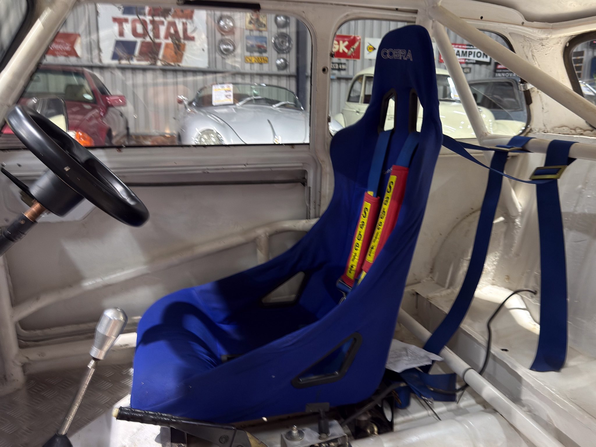 Interior of a race car cockpit with a blue racing seat and exposed roll cage in a workshop setting outside cars on display.