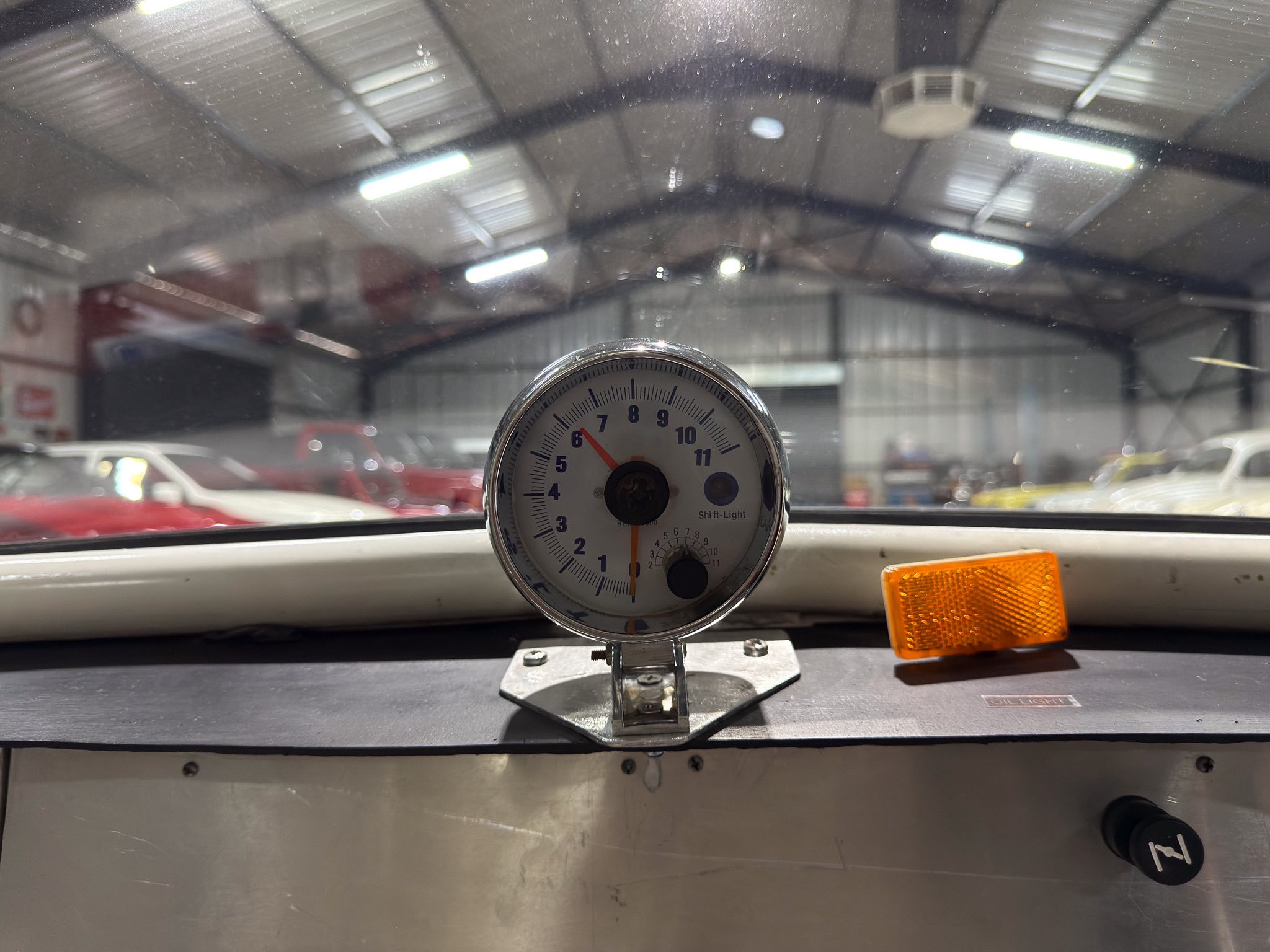 Circular analog gauge mounted on a car dashboard in a workshop, with an orange reflective marker to the right and a blurred garage interior in the background.