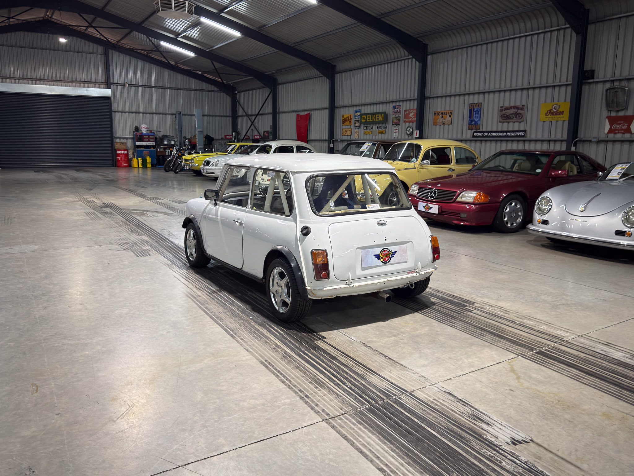 Indoor garage with rows of vintage cars on display in a metal-walled warehouse park, white car in foreground and classic cars along wall
