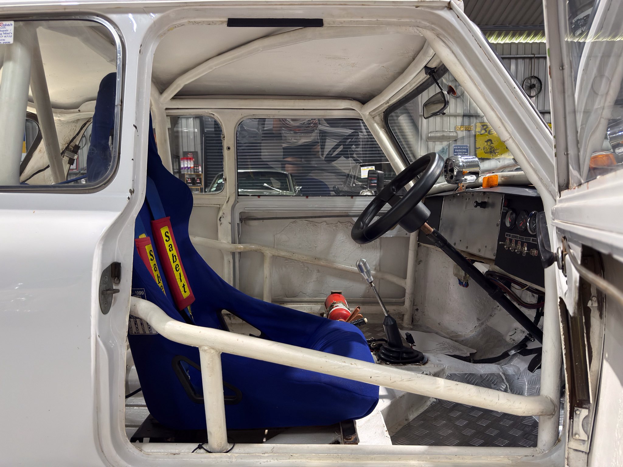Interior of a white race car showing a blue racing seat with yellow Sabelt harnesses and a steering wheel in a workshop environment; roll cage and metal dash visible.