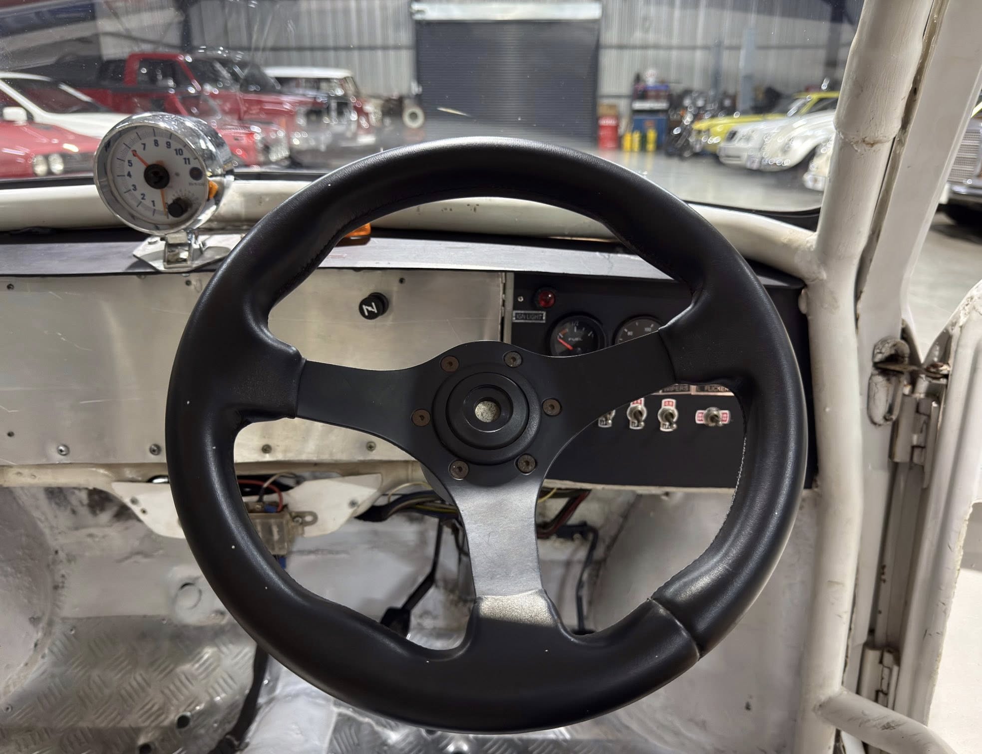 Close-up of a black steering wheel inside a stripped car cockpit, with gauges and switches on a metal dashboard and a workshop full of vintage cars in the background.
