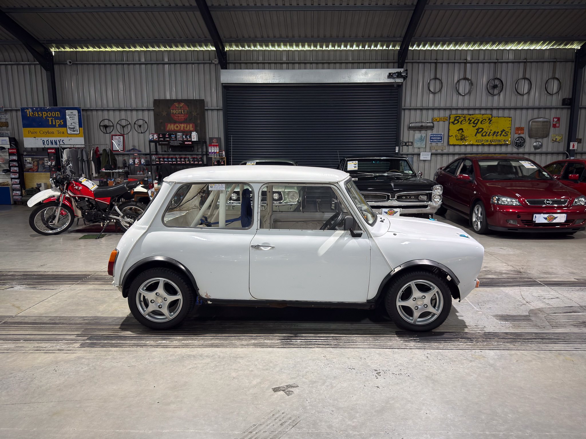 Inside a car showroom with vintage vehicles: a white classic car in the foreground and a red motorcycle on the left, metal walls, and classic signs in the background.