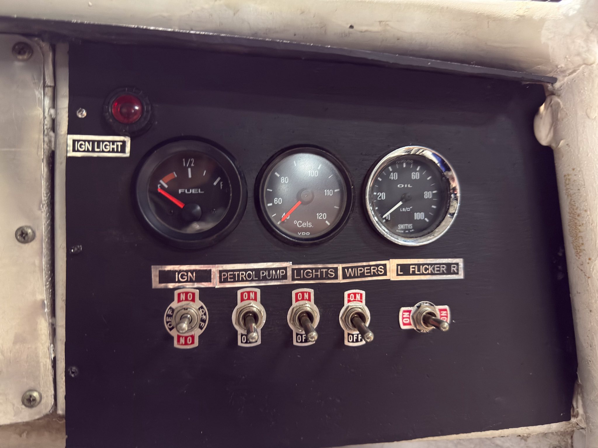 Close-up of a vintage control panel with four round gauges (fuel, temperature, oil) and a row of ON/OFF switches under labels like IGN LIGHT and LIGHTS on a dark dashboard.