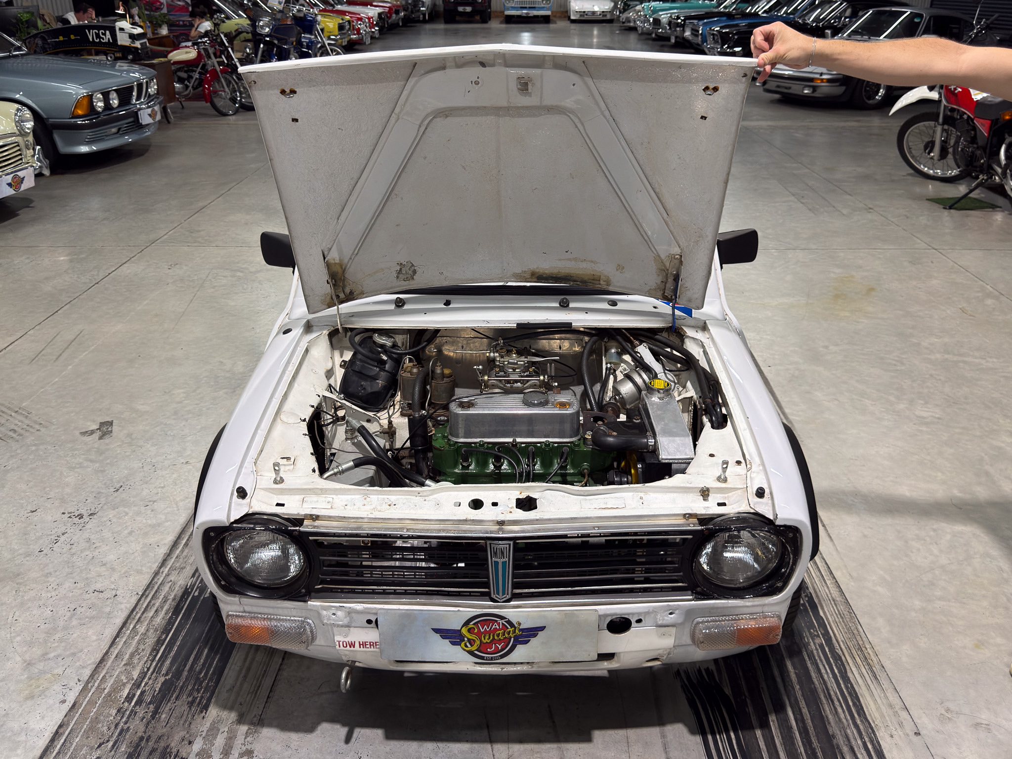 White classic car with its hood raised, exposing the engine in a showroom filled with vintage cars; a person’s arm holds the hood edge to the right.