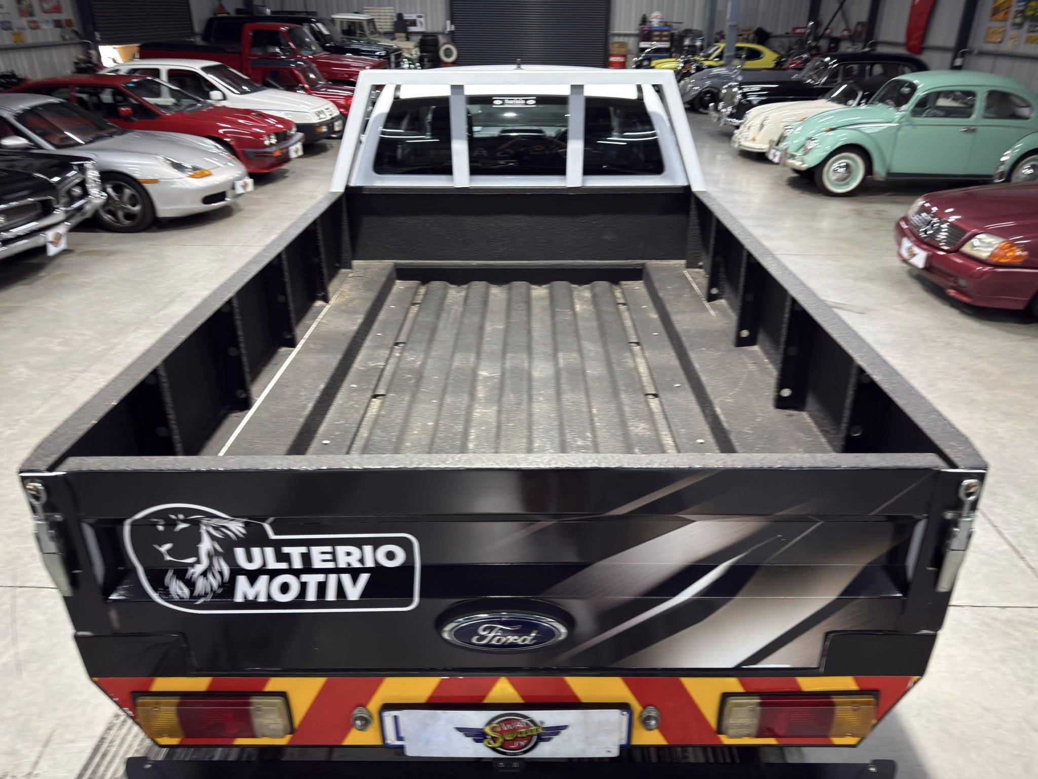 Rear view of a black Ford utility trailer in a showroom, surrounded by vintage and modern cars on both sides.