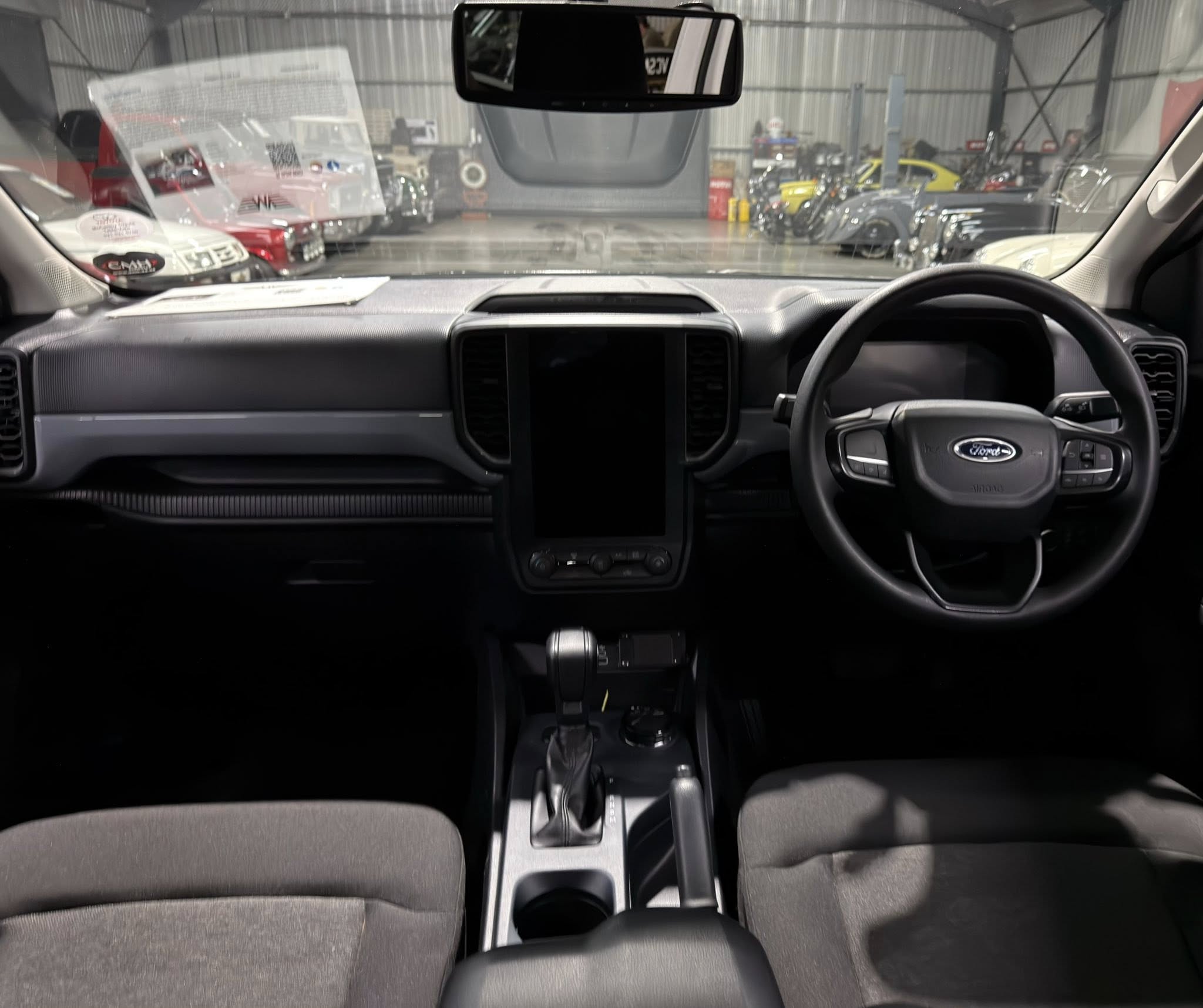 Driver's-seat view of a Ford, showing the dashboard, center console and gear shifter; showroom with multiple cars visible through the windshield.
