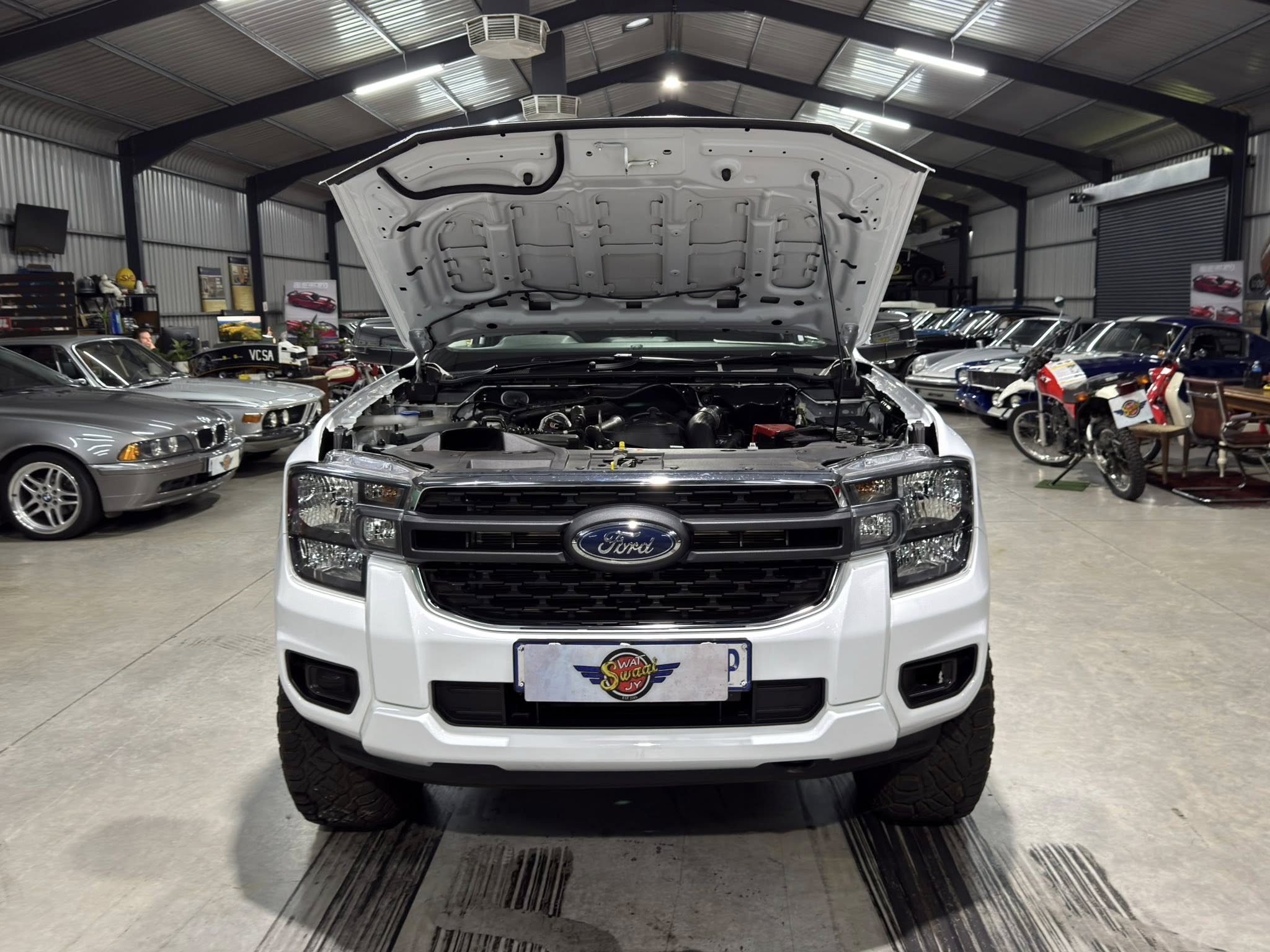 White Ford truck with its hood open in a showroom or workshop, exposing the engine bay amid other cars and motorcycles in the background.