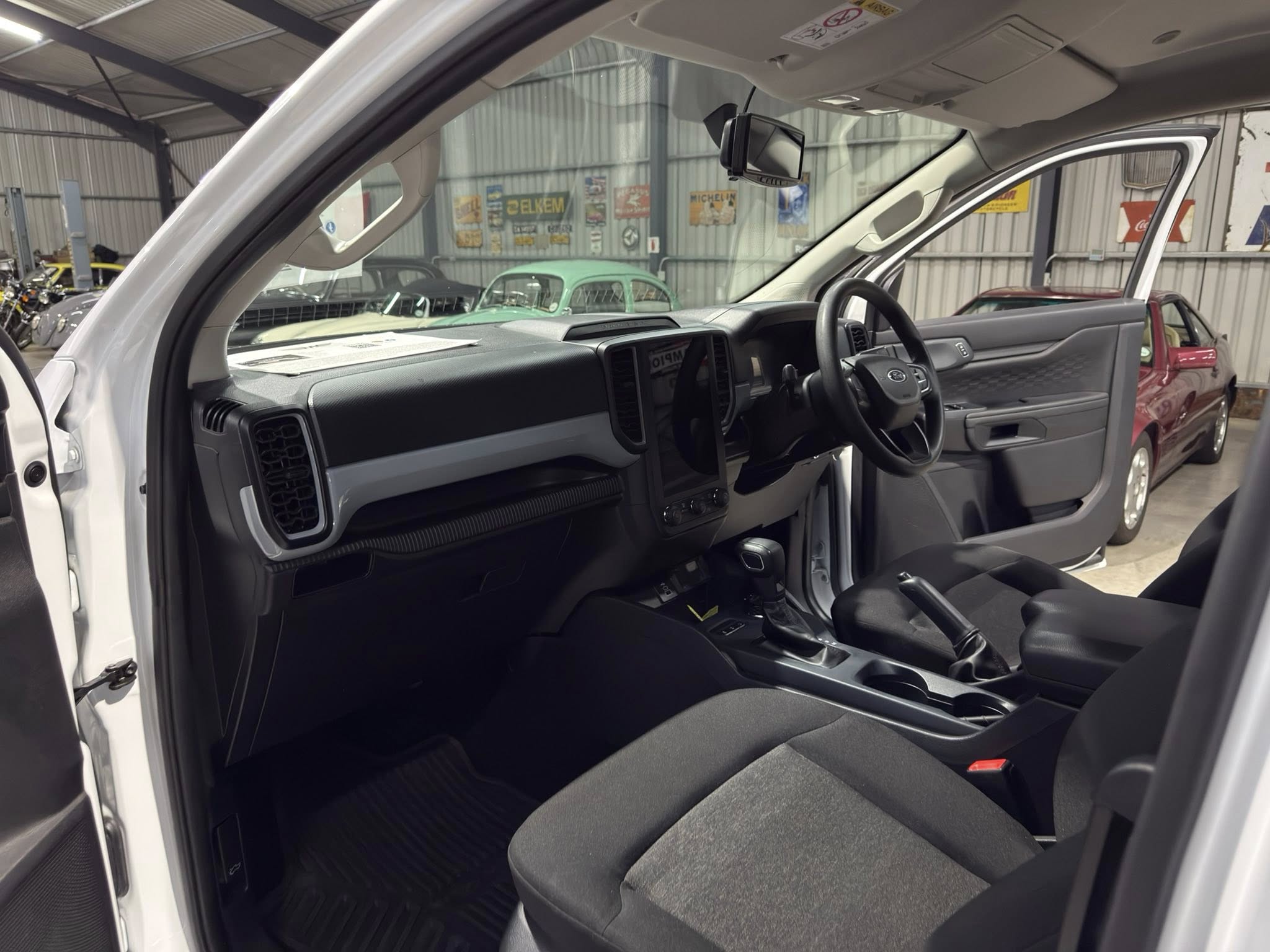Driver-side view of a white pickup truck interior with dashboard, steering wheel, and center console visible inside a showroom showroom filled with other color cars in the background.
