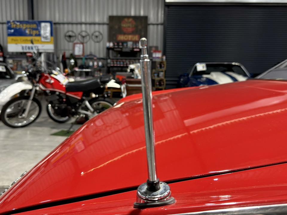 Close-up of a chrome antenna mounted on the red hood of a classic car, with motorcycles and workshop items in the background.