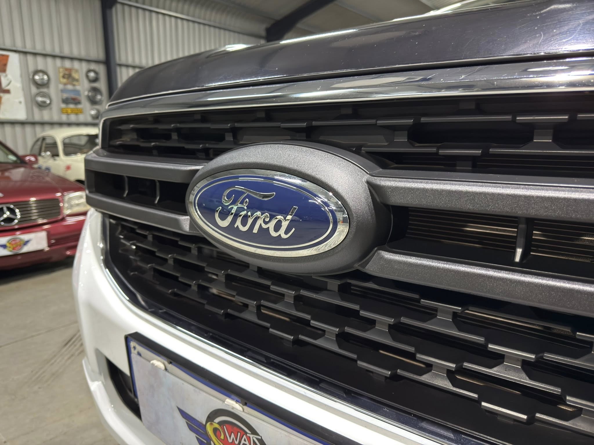 Front grille of a Ford vehicle with blue oval badge, chrome trim, indoors in a workshop with other classic cars in the background.