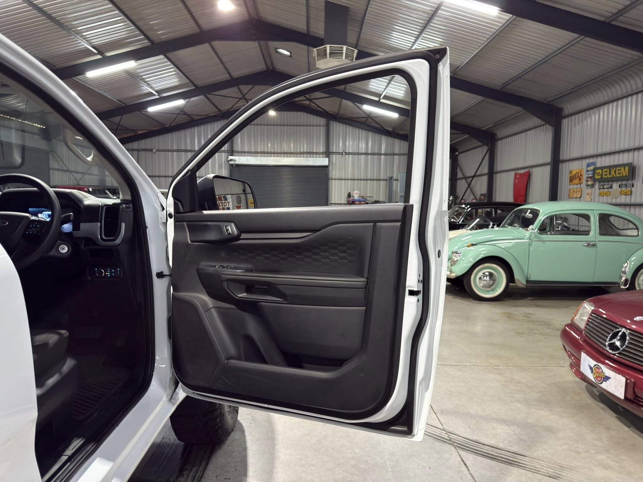 Open front passenger door of a white pickup inside a showroom or garage, with vintage cars and retro signs visible in the background.