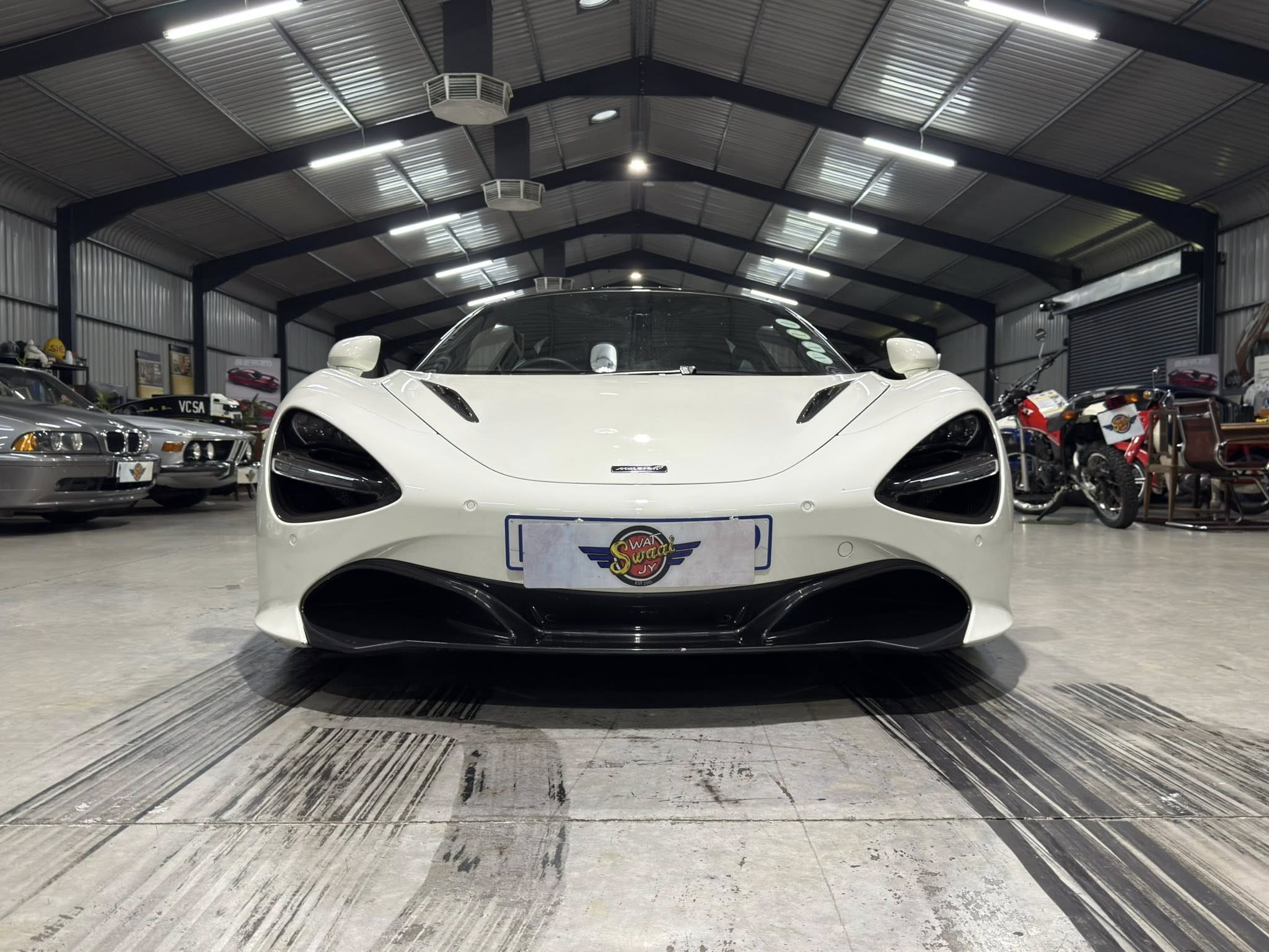 Front view of a white sports car in a showroom, shot from ground level with other cars and bikes in the background.