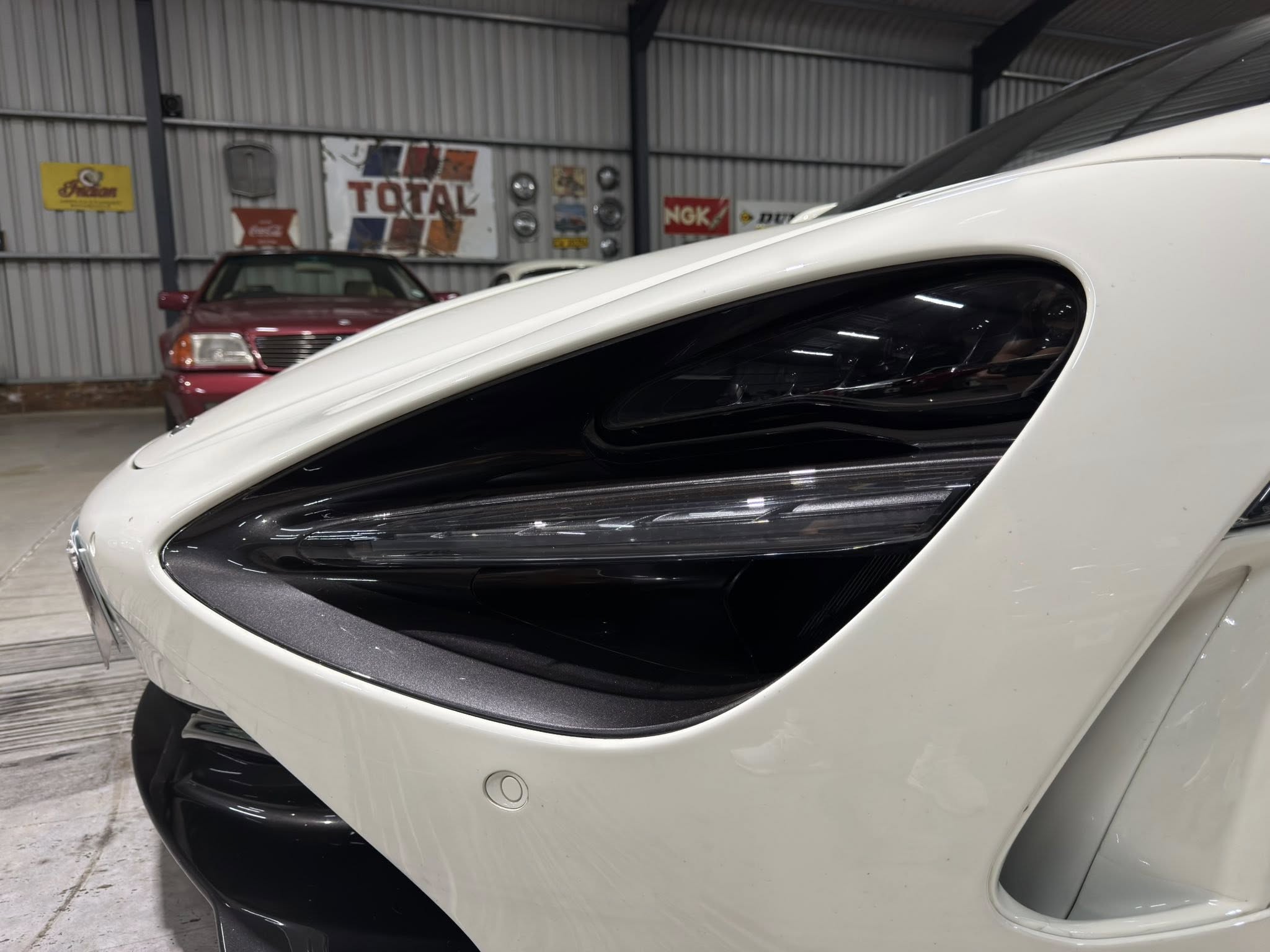 Close-up of a white car’s front with a curved windshield, inside a metal-wall garage with vintage signs in the background.