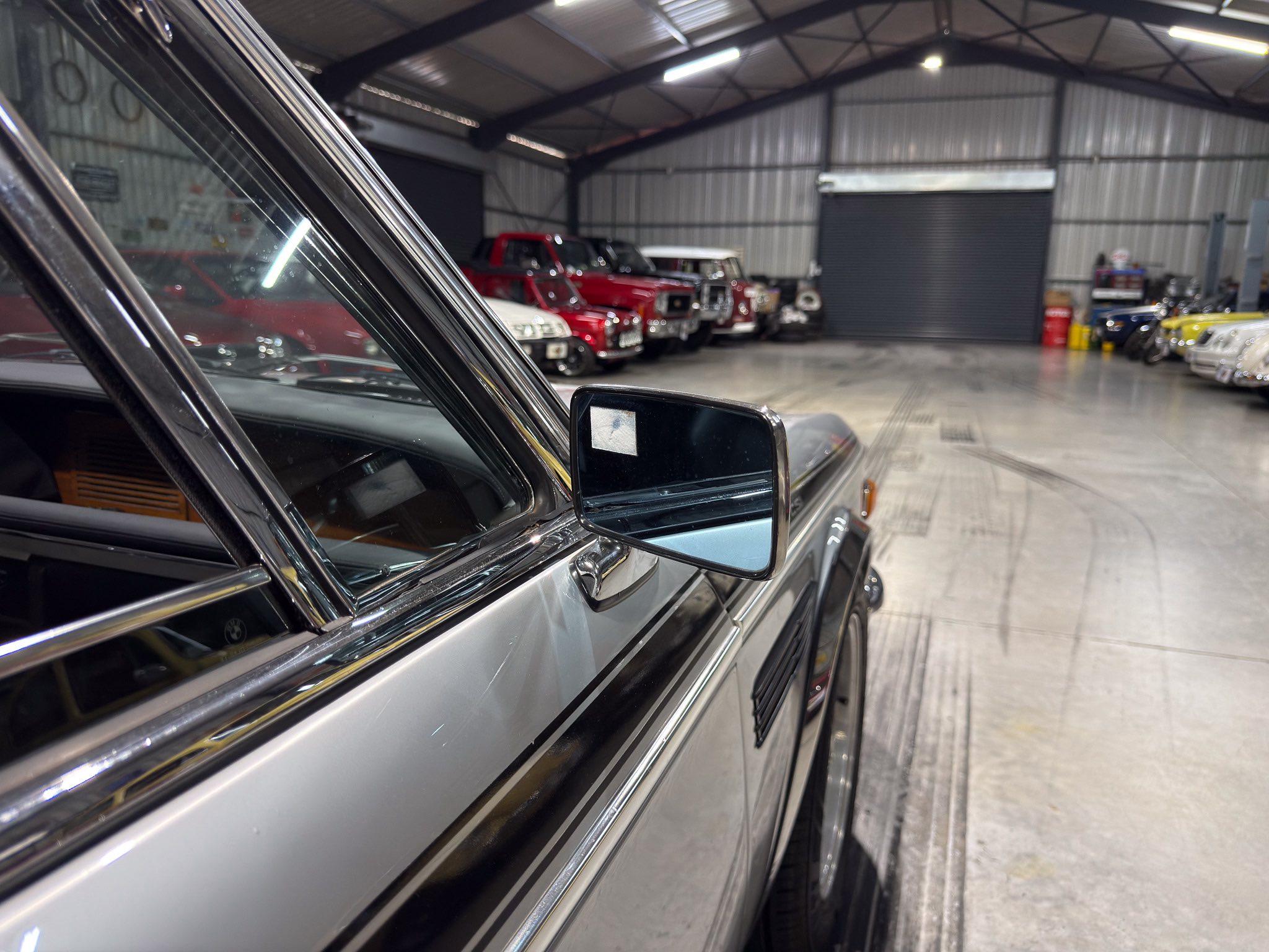 Close-up of a silver car door and side mirror in a large indoor garage with vintage cars lined up along the walls.
