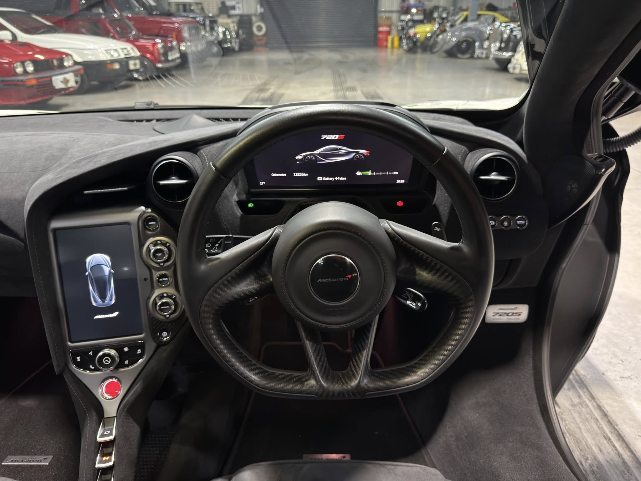 Interior view of a McLaren cockpit with a steering wheel and a central digital instrument cluster showing a car illustration and performance readouts, with a showroom of parked cars visible through the windshield.