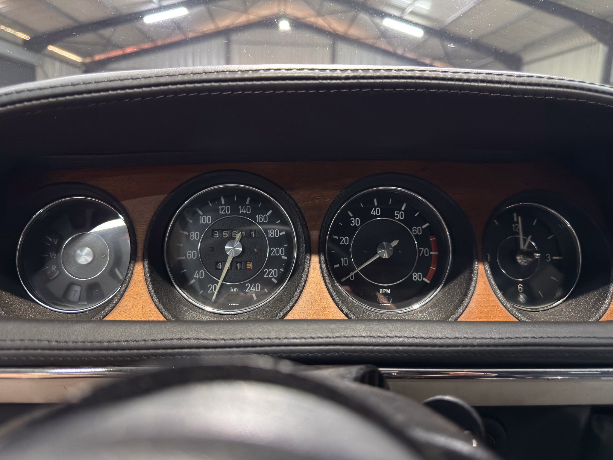 Close-up of a classic car dashboard with analog gauges (speedometer center) and wood trim, stitched black dash, viewed from the driver’s perspective with the top of the windshield area visible above.