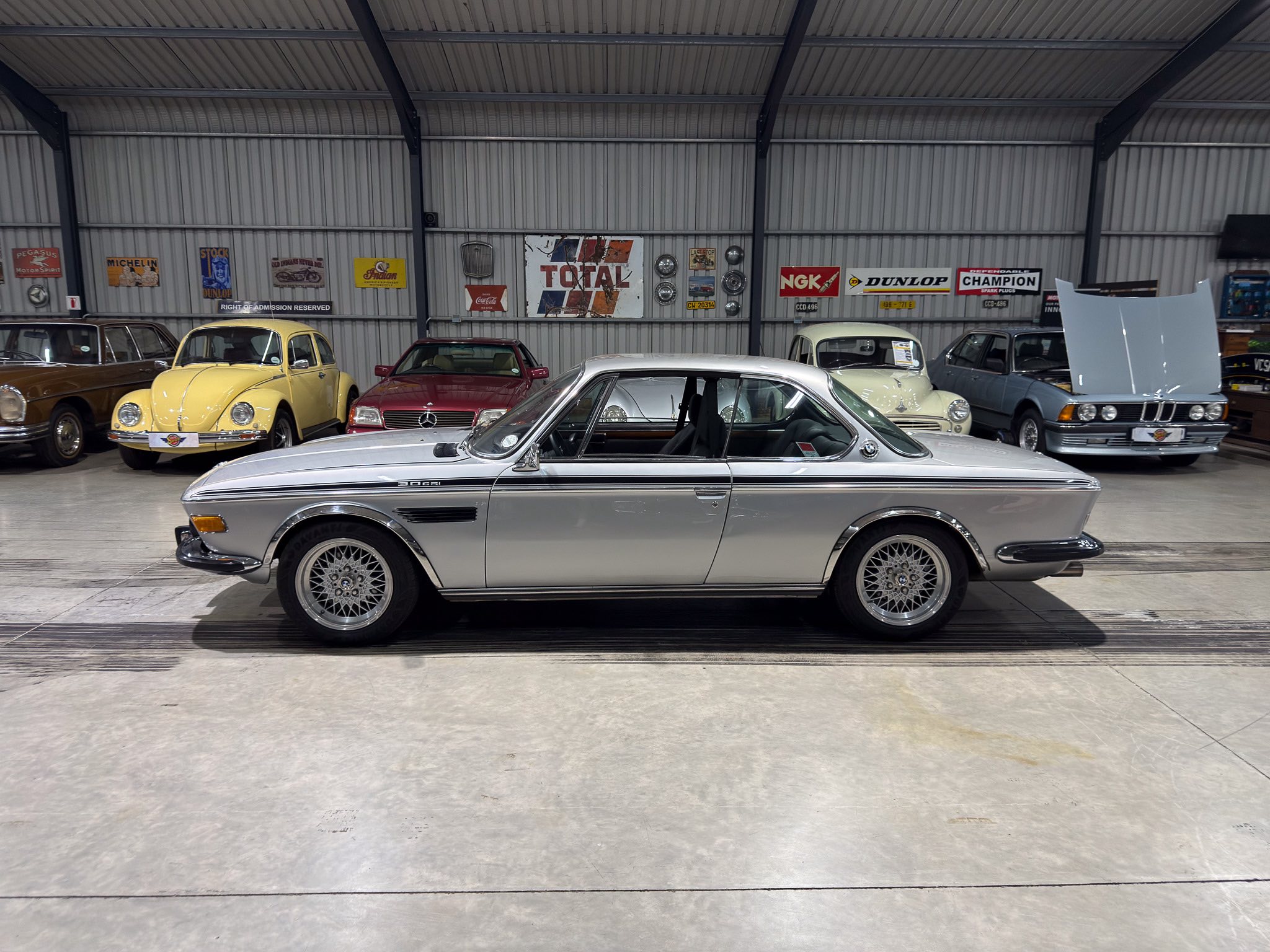 Indoor showroom with several vintage cars, a silver car in the foreground and a yellow car behind, against a wall of retro signs and a metal garage interior.