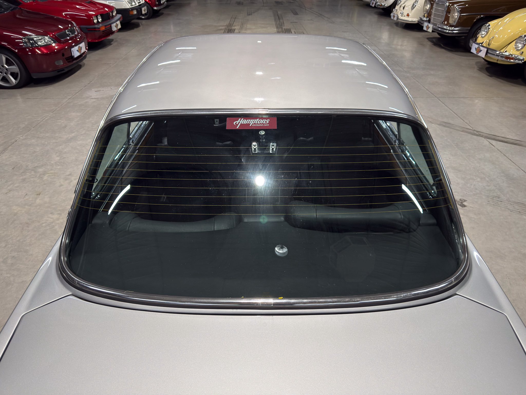 Back view of a silver car in a showroom, with rows of colorful classic cars on both sides and a Hamptons sticker on the rear window.