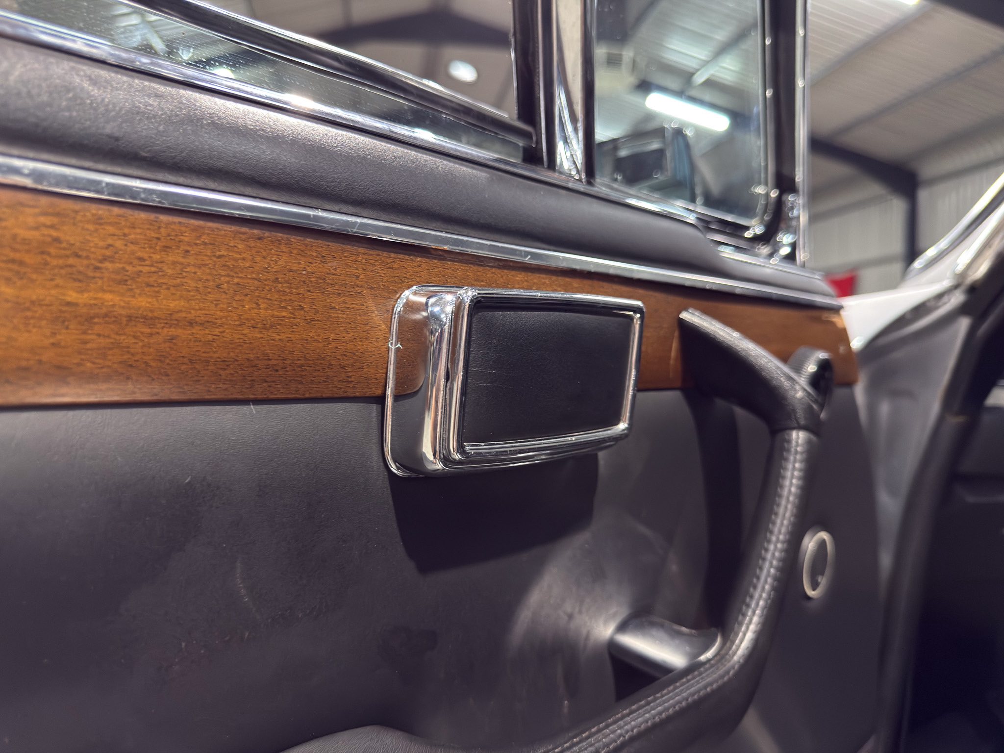Close-up of a car interior door panel with dark leather, wood veneer trim and chrome accents; window frame visible in the background.