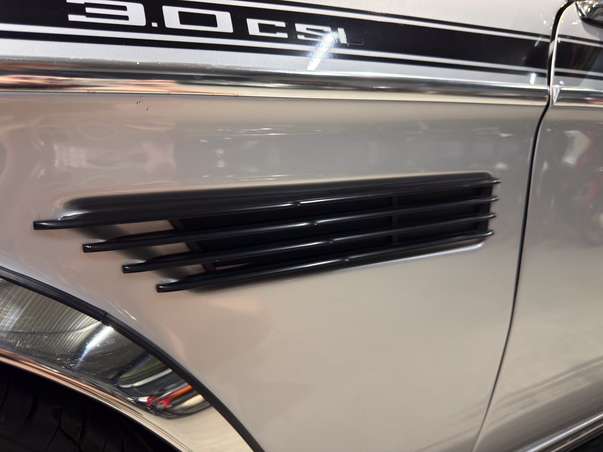 Close-up of a classic car door with horizontal chrome trim, vent-like louvers, and a partial headlight at the bottom left corner.