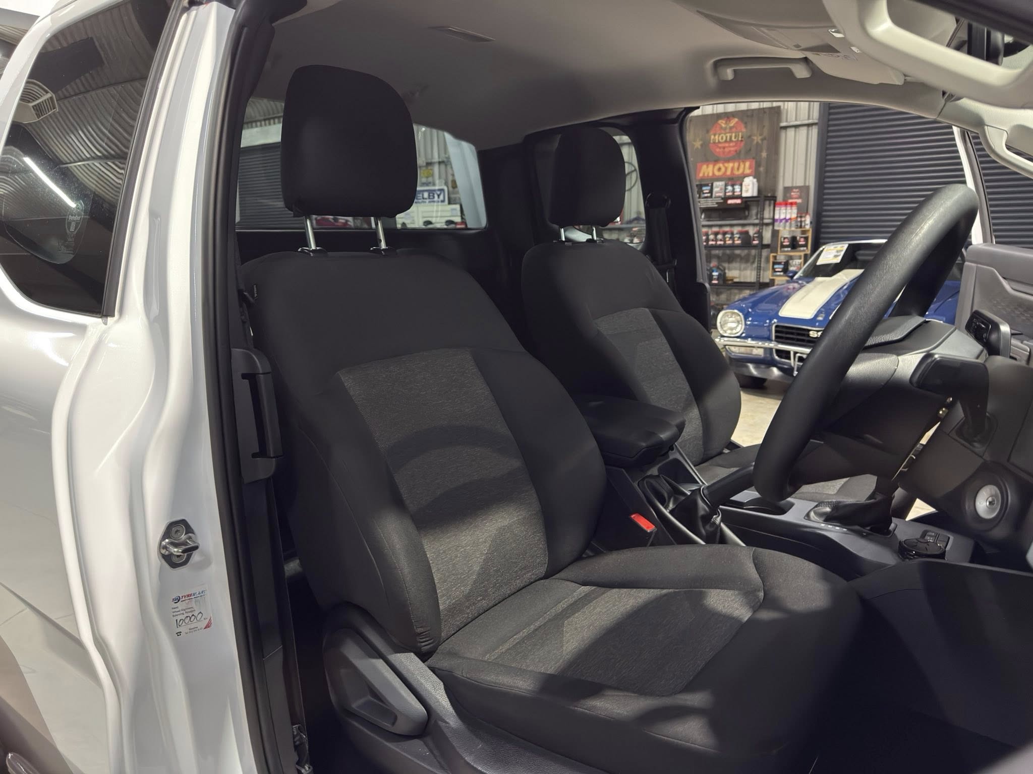 Inside a white SUV showing the driver and front passenger seats with black upholstery; the driver's door is open in a showroom, with another blue car visible in the background.