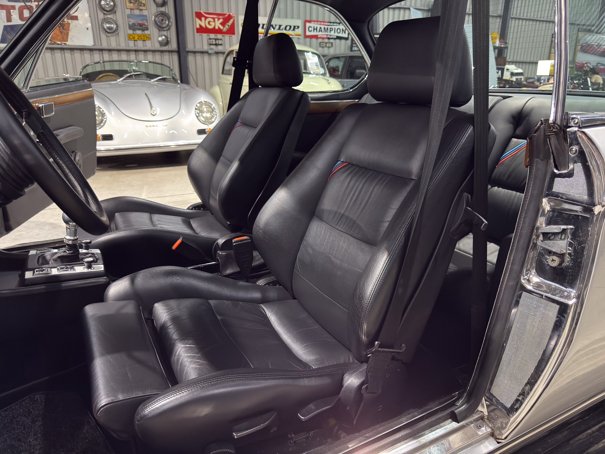 Front interior of a classic car with black leather seats, open door, and seat belts visible in a showroom background with vintage signs and other cars.