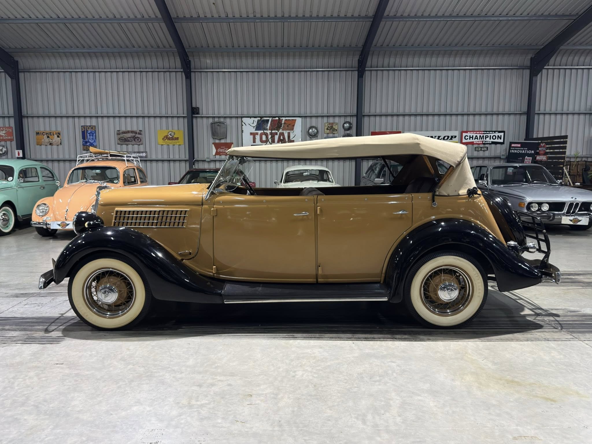 Bronze vintage convertible with its beige roof partially up, parked among other classic cars inside a retro showroom with metal walls and signage on the back wall.