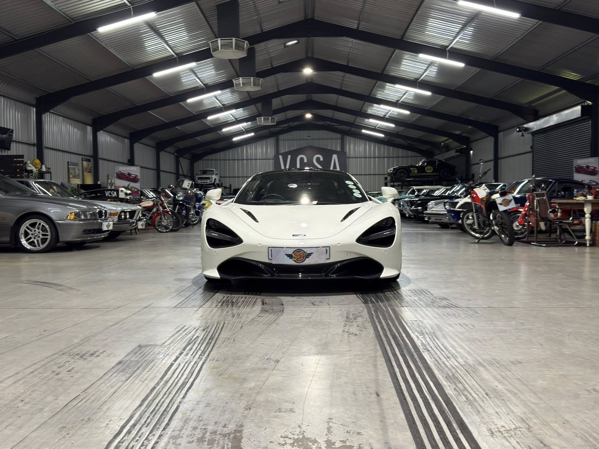 White sports car centered in a large showroom with other cars and motorcycles along the sides; VCSA sign in back.