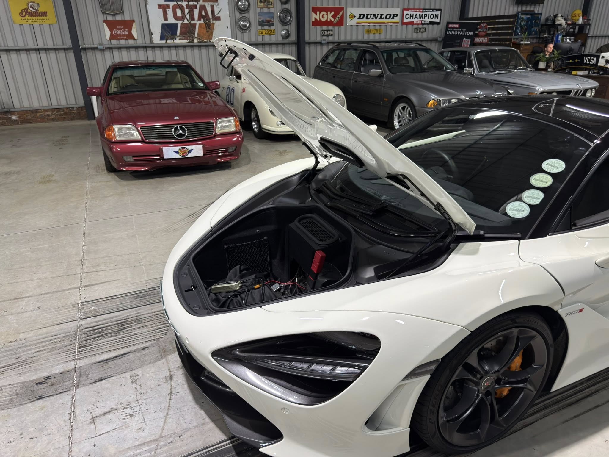 White sports car with its hood raised in a showroom/garage, surrounded by vintage cars and retro signs on the walls.