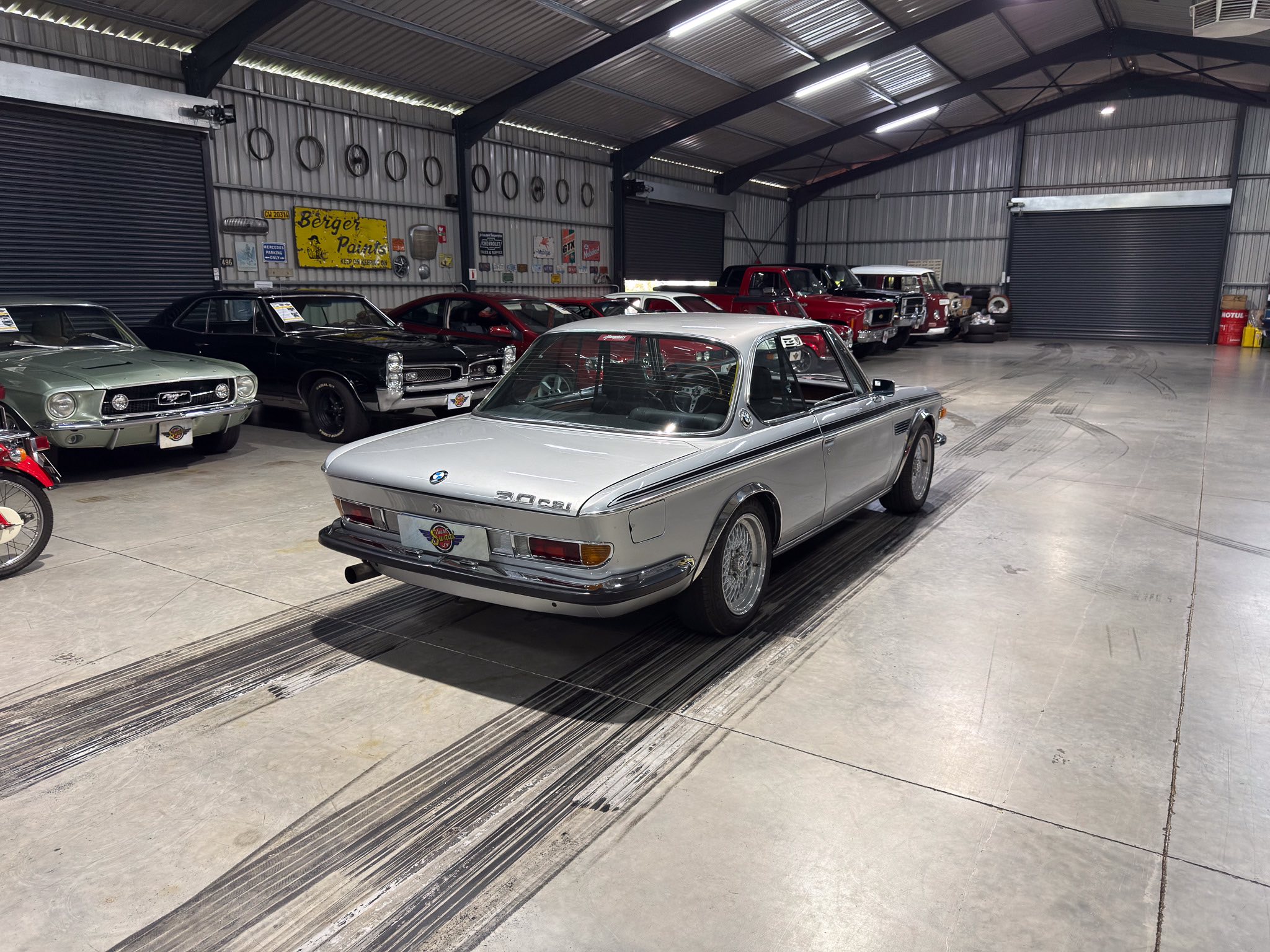 Silver BMW 3.0 CSL in the foreground of a classic car showroom with a row of vintage cars behind it on a polished concrete floor.