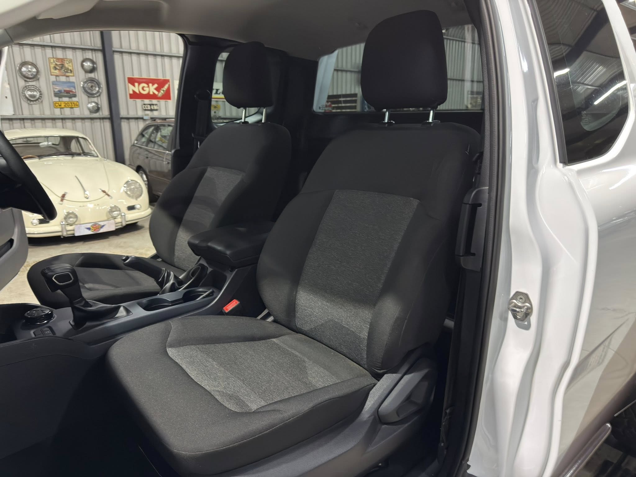 Front interior of a car showing two front seats, center console and gear lever with a showroom of vintage cars in the background through an open door window.