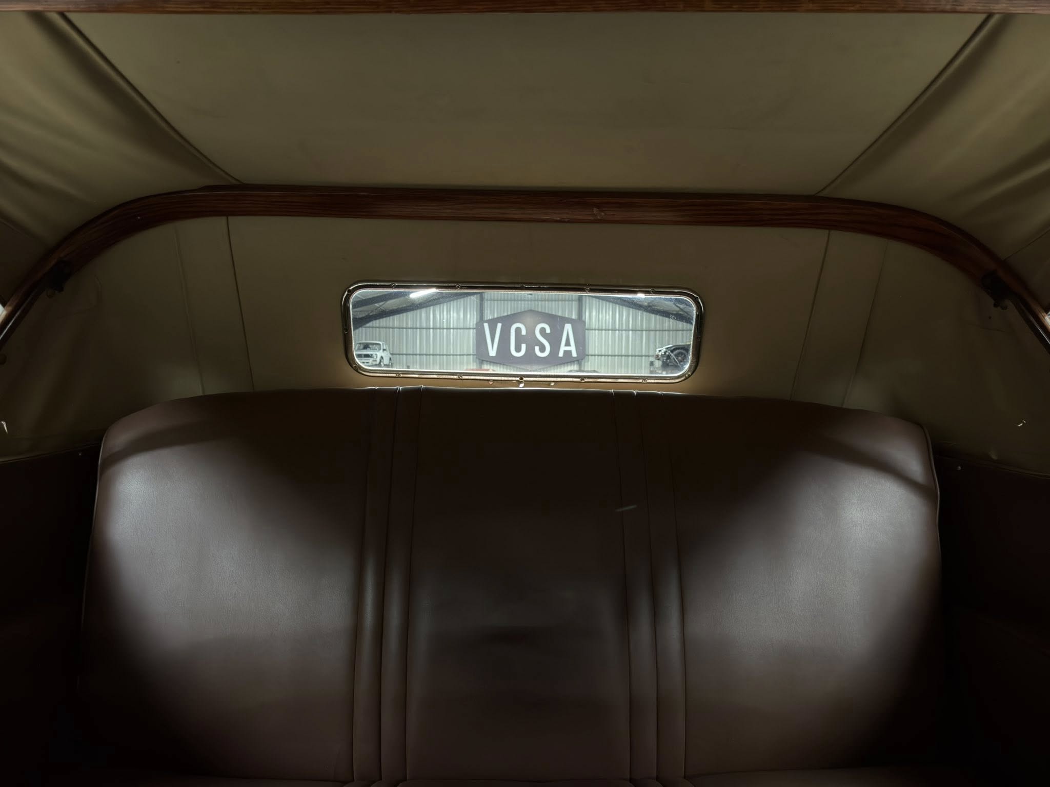 Back seat of a vintage car with brown leather and wood trim; rear window shows a building and a 'VCSA' sign.