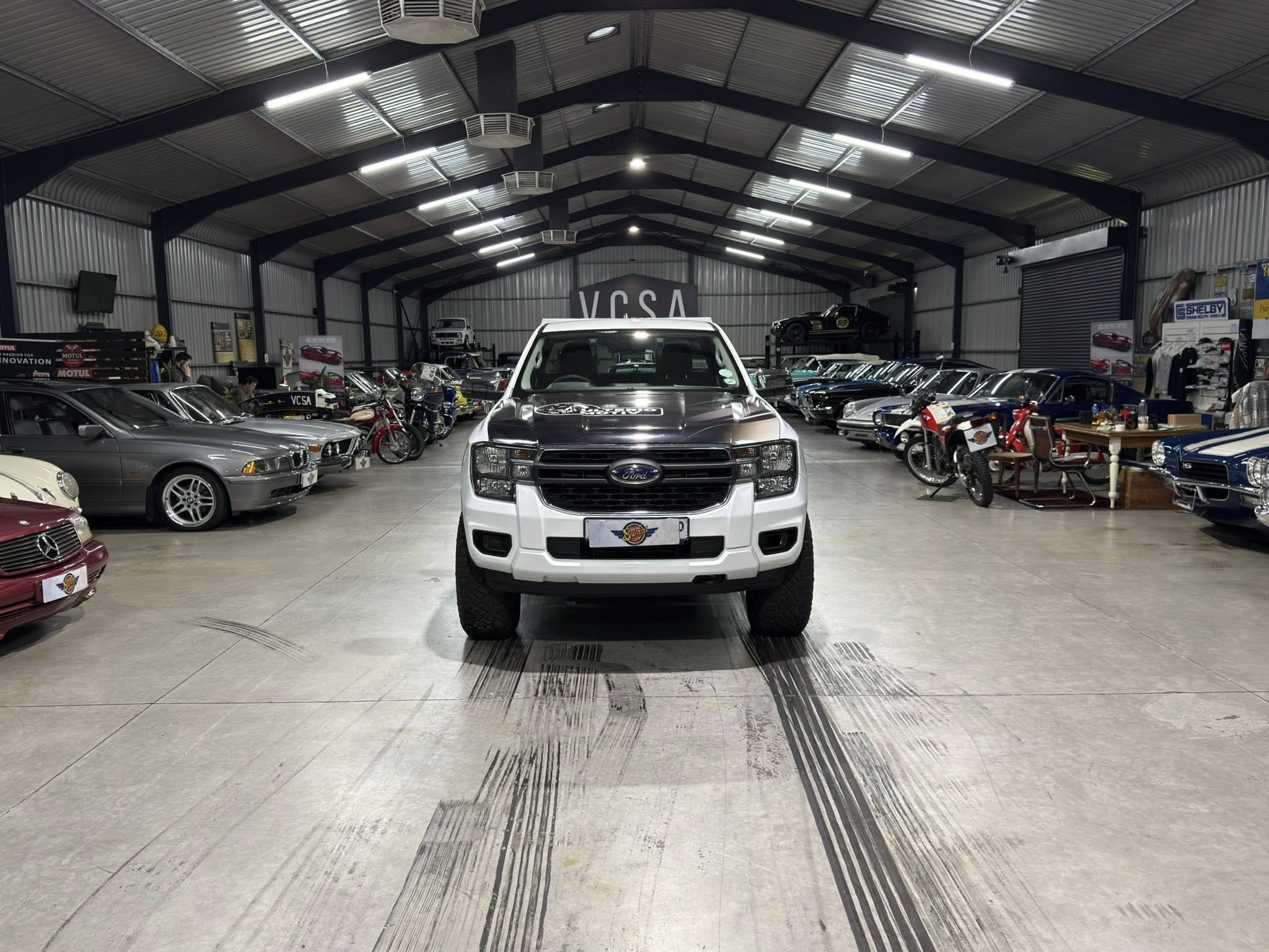 White pickup truck centered in a large indoor car showroom with many cars and motorcycles on both sides.
