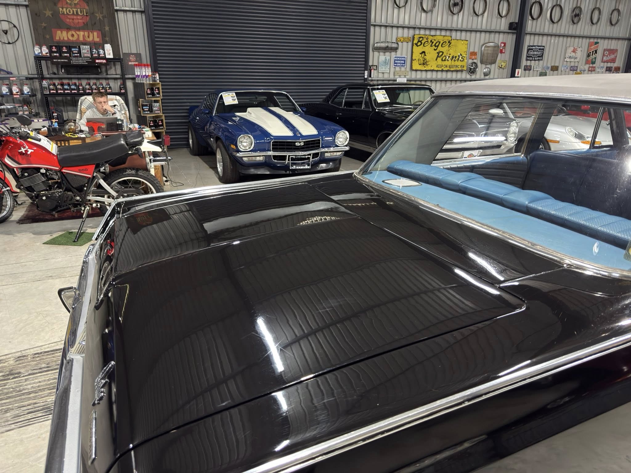 Inside a garage or showroom with classic cars: a blue 1969 Camaro SS with white racing stripes center, others parked around, and a man at a desk on the left.